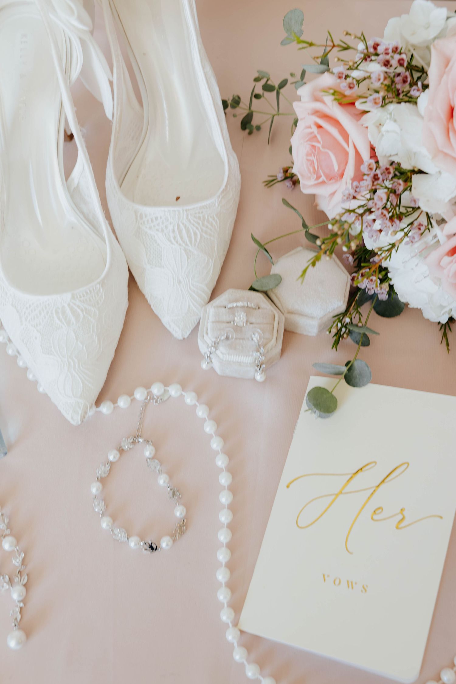 Bridal flat lay with white lace heels, pearl jewelry, vow book, ring box, and pink floral bouquet on blush background in Lansing Michigan.