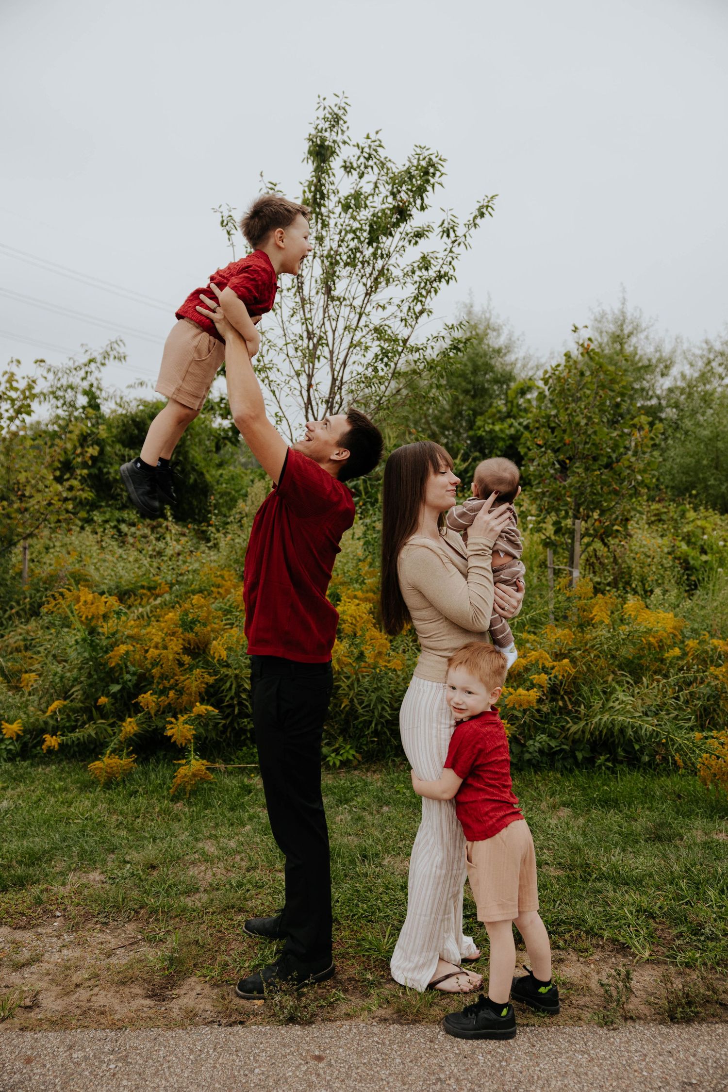 Playful outdoor family photography session featuring parents lifting their children into the air in a grassy field surrounded by trees.