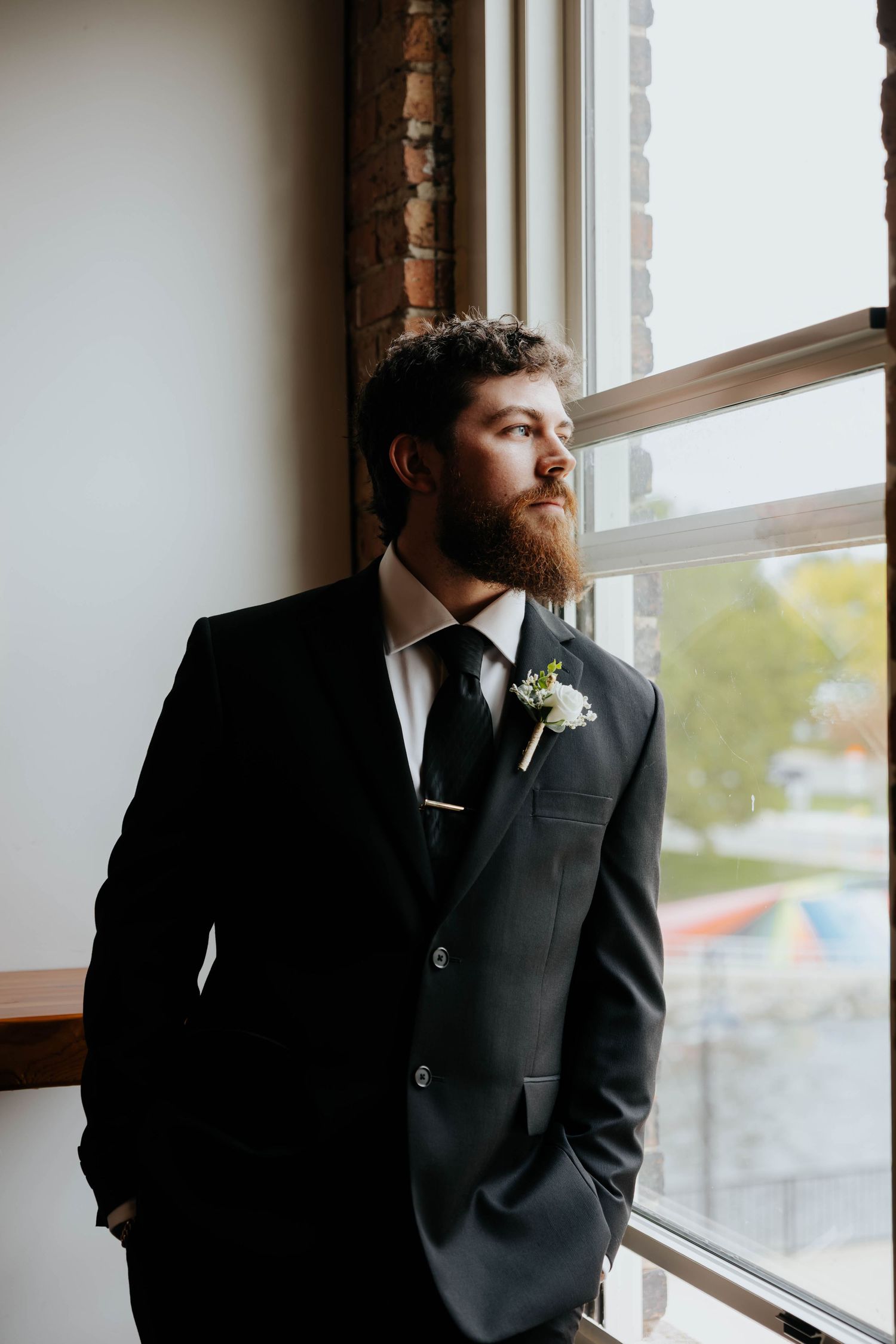 Classic groom portrait in a black suit beside a window at Record Box Loft, a wedding venue in Battle Creek, Michigan.
