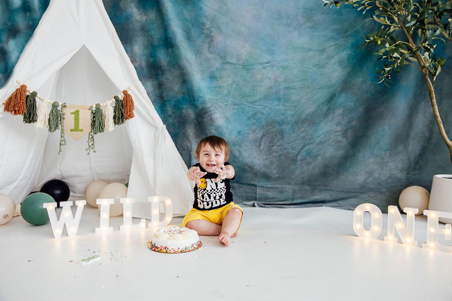 Wild One themed first birthday cake smash photo of a baby boy sitting beside his birthday cake with a teepee and blue backdrop.