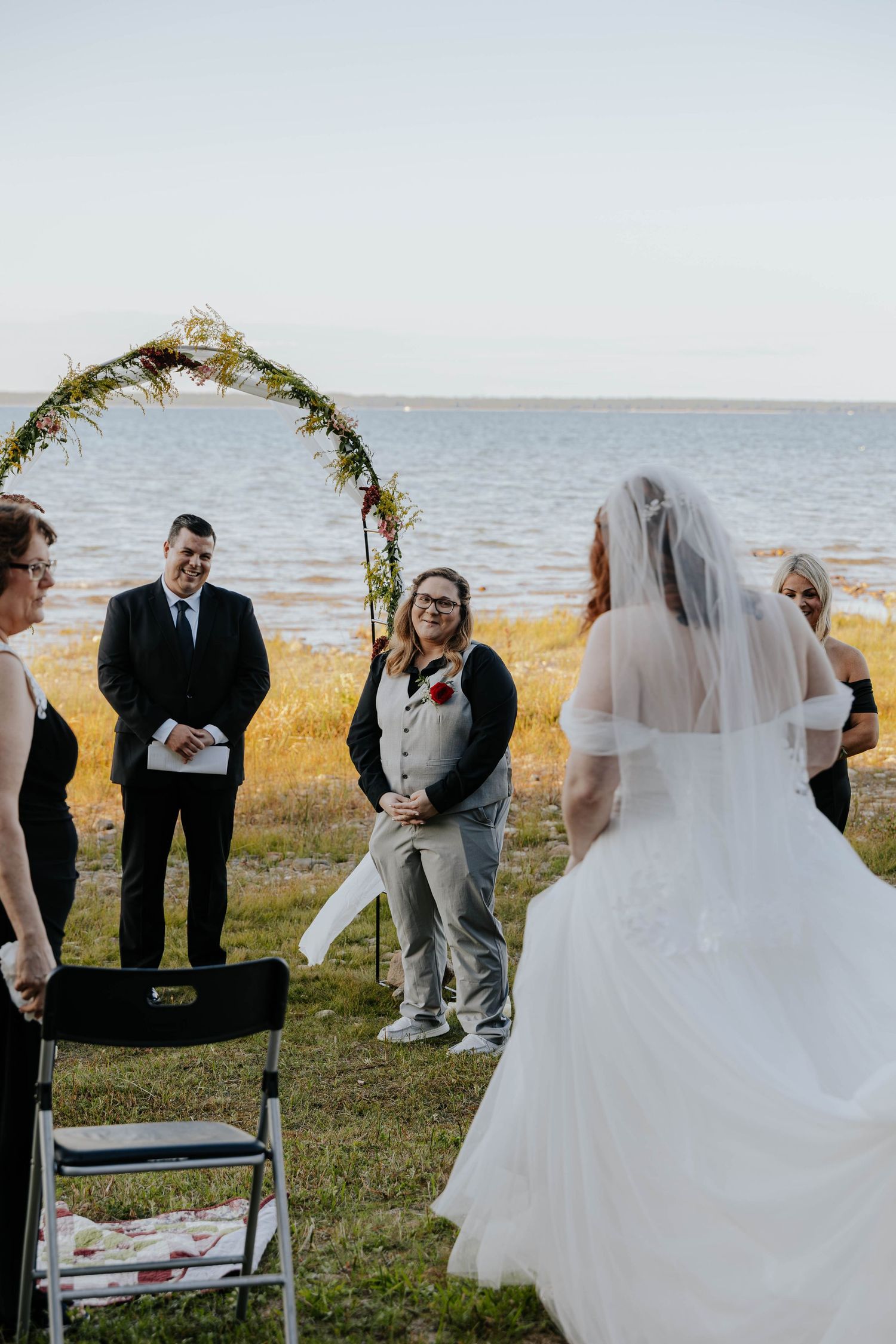 Bride in a flowing white wedding gown approaching a floral ceremony arch overlooking the water at a Northern Michigan elopement.