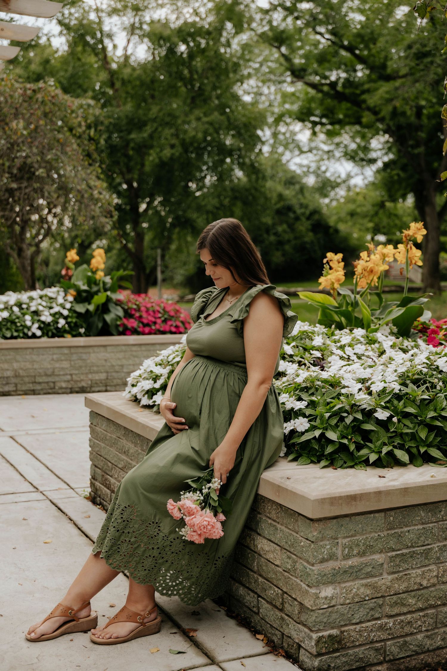 Outdoor maternity portrait of an expecting mother in a green dress sitting beside a flower garden in Lansing, Michigan.