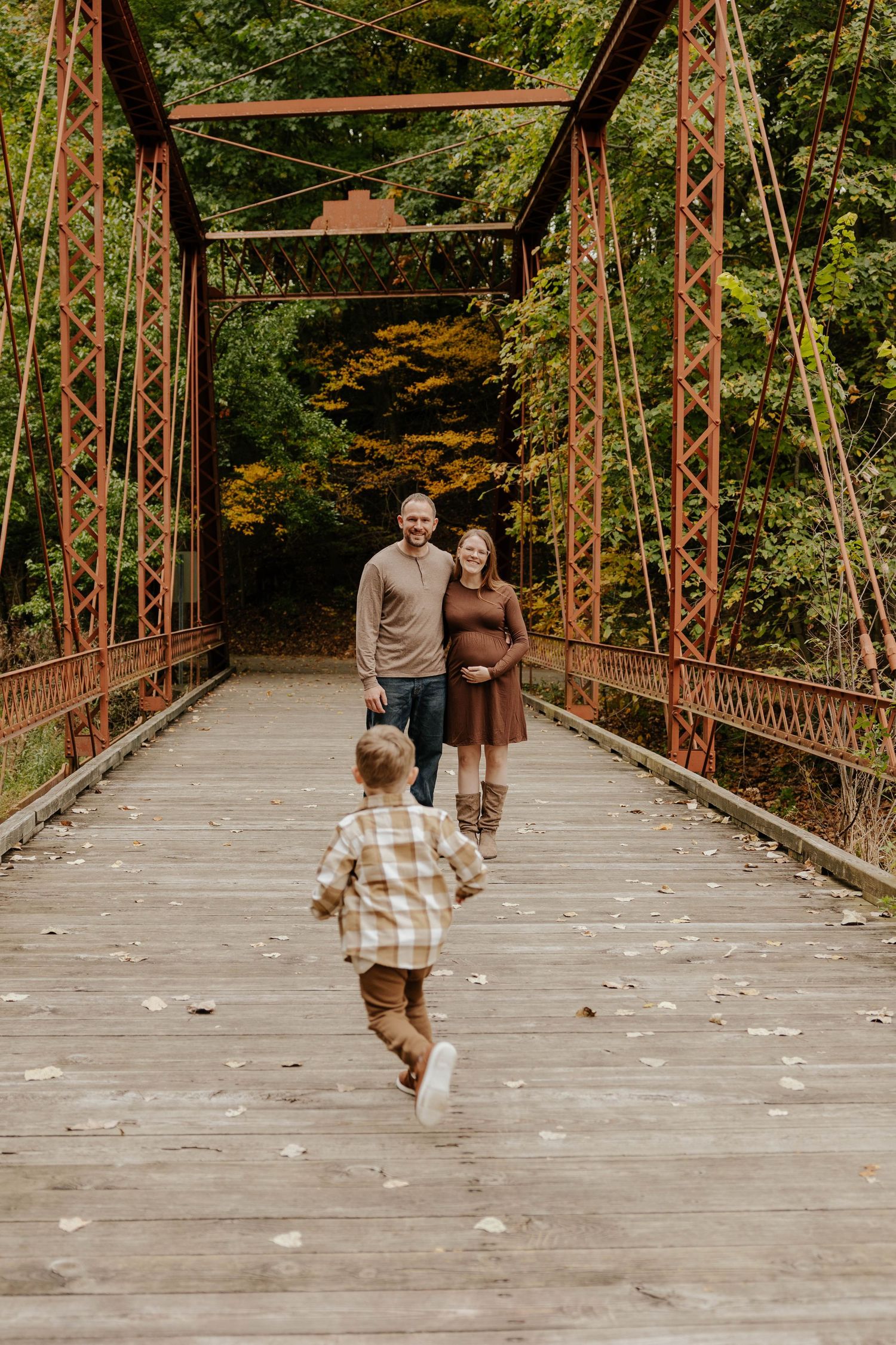 Candid fall family photo of a young child running across a rustic bridge surrounded by colorful autumn foliage.