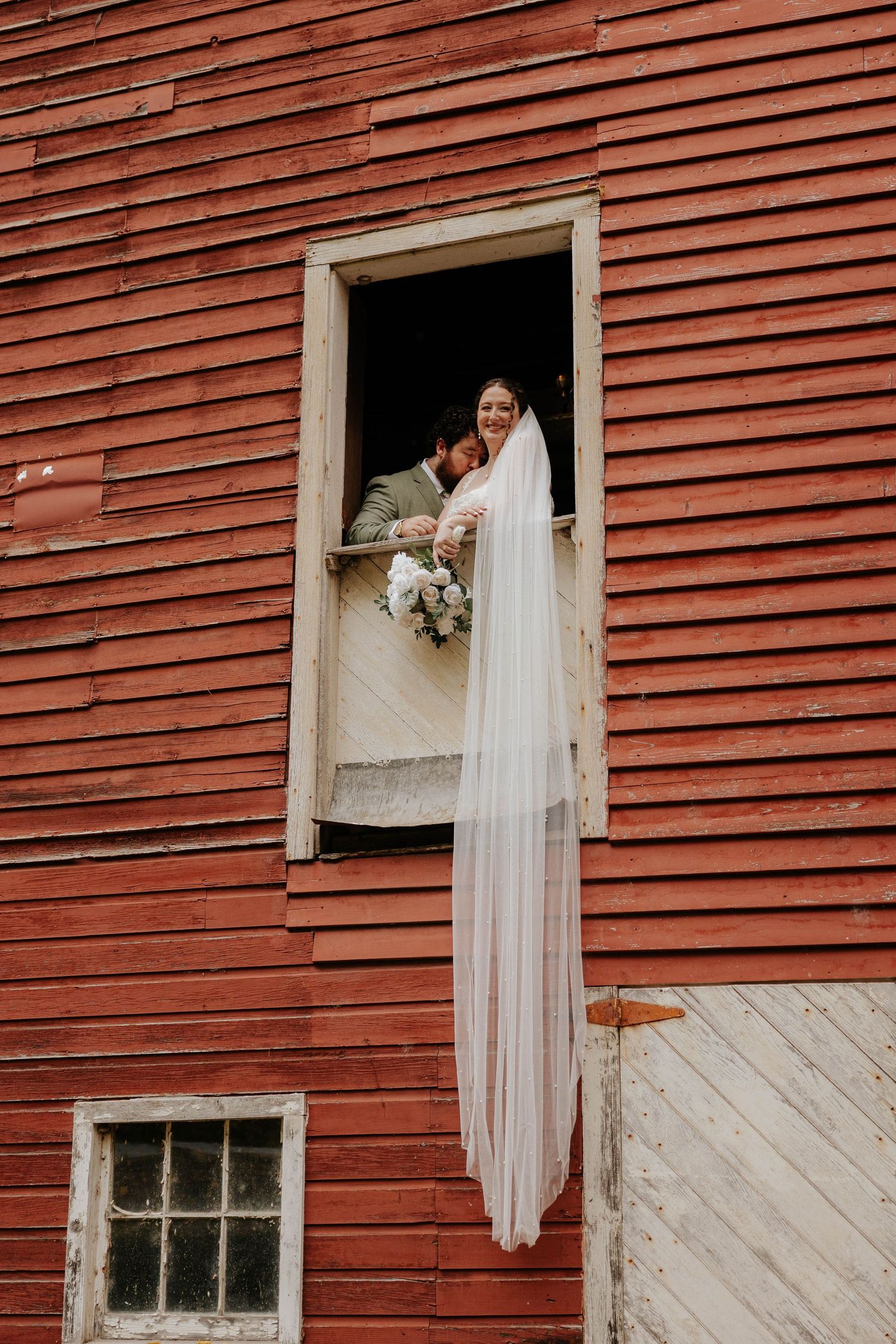 Creative wedding portrait of a bride standing in a rustic red barn window at Bowen Mills in Middleville, Michigan.