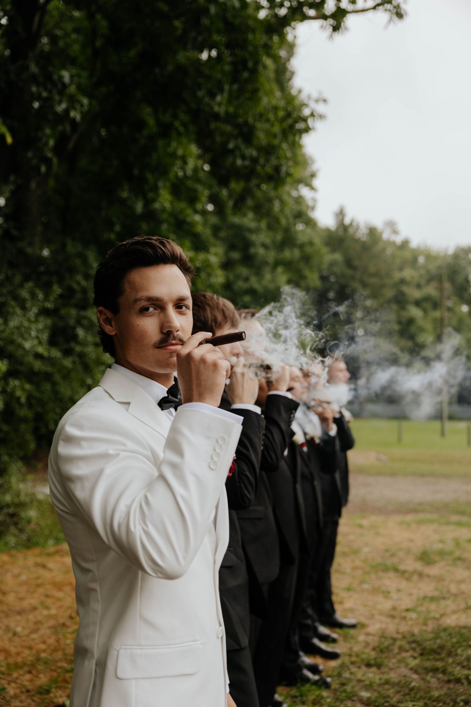 Groom in white suit smoking cigar with groomsmen in black suits lined up outdoors at a wedding celebration.