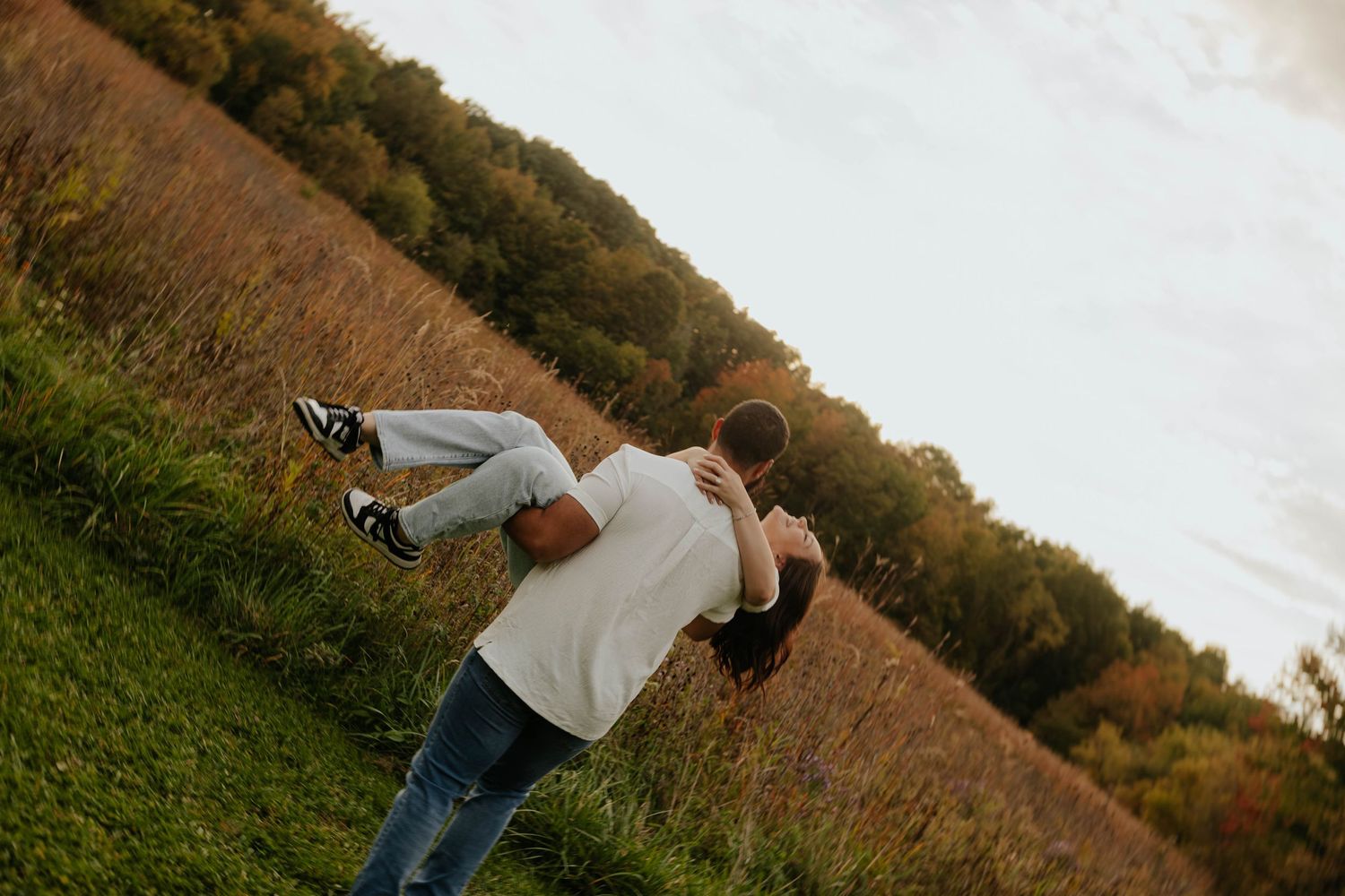 Playful engagement photo of a man lifting his partner in a grassy field during a romantic fall couples session in Caledonia, Michigan.