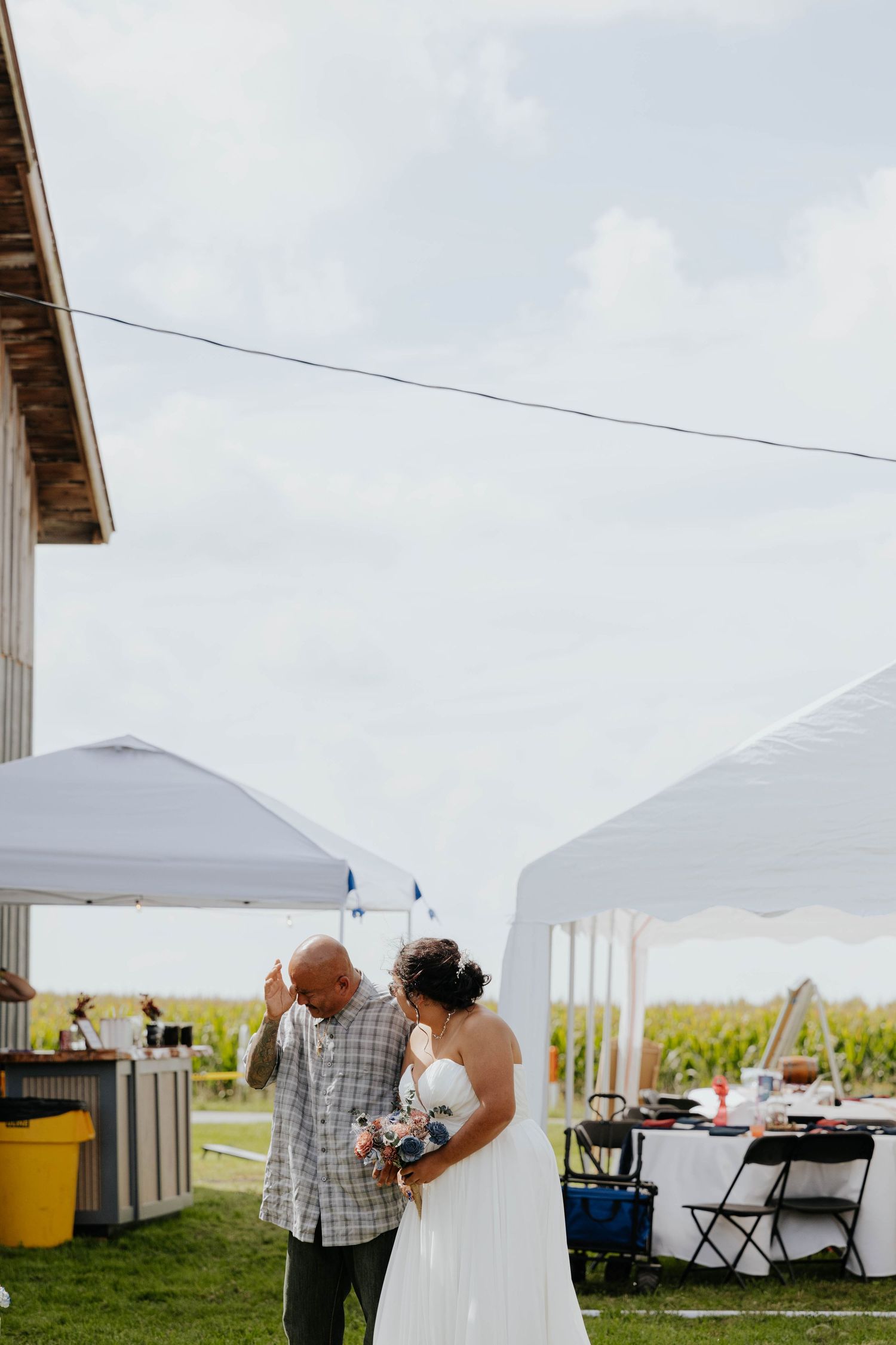 Bride in white dress holds bouquet while sharing a moment with a guest at an outdoor farm wedding reception.