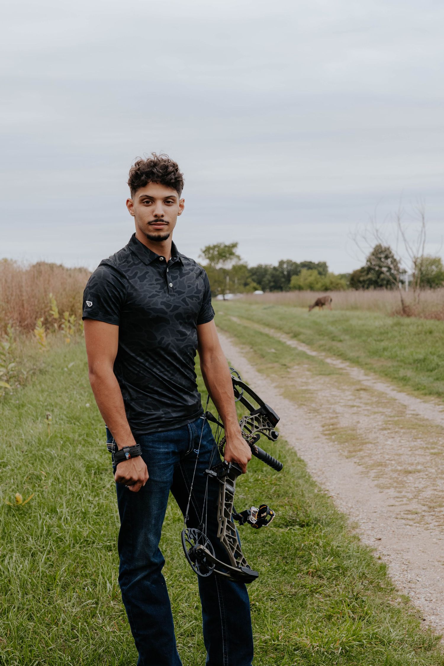 Outdoor senior portrait of a high school senior wearing a dark shirt and jeans, standing beside a country road and grassy field.