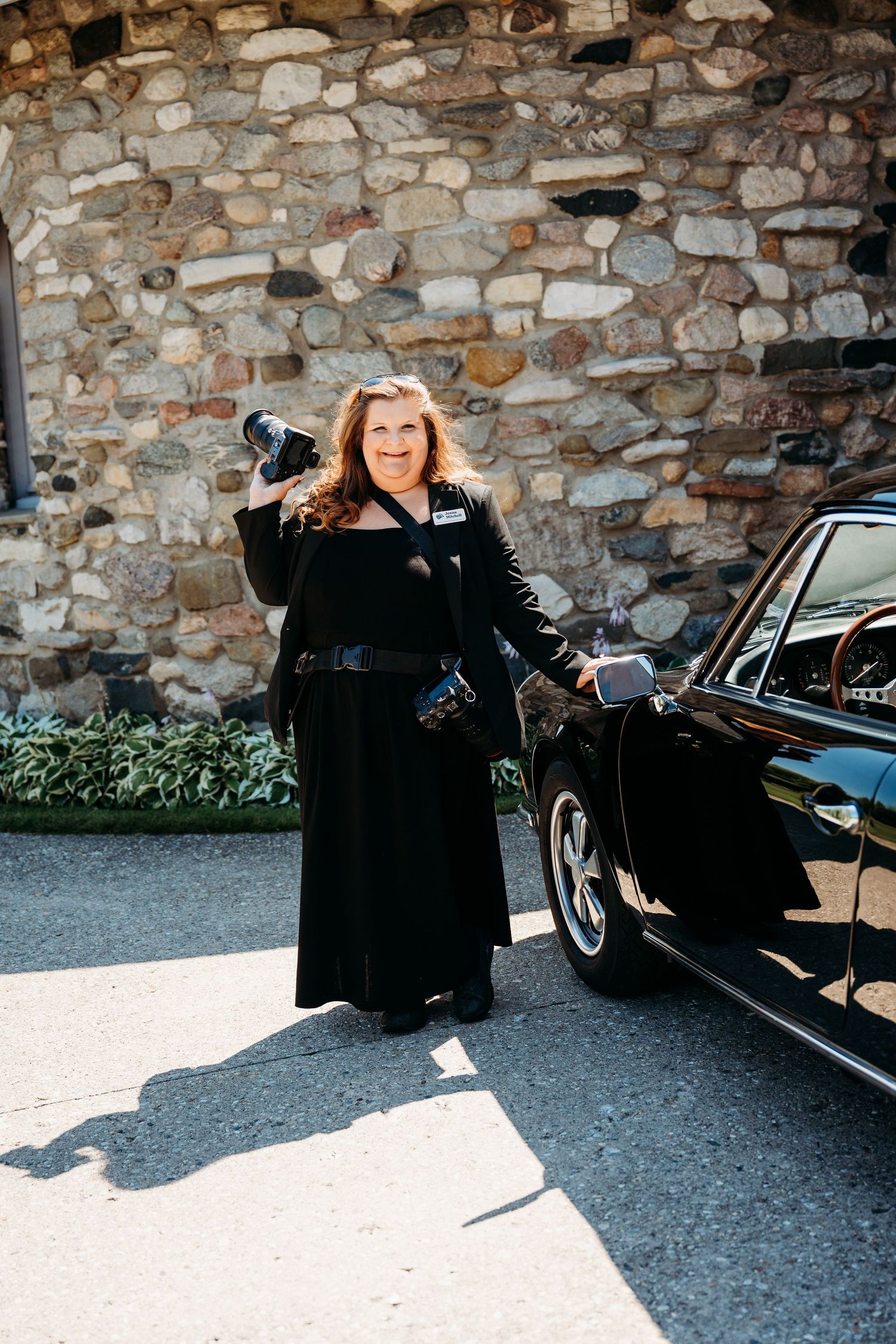 Woman in an elegant black outfit poses beside a stone wall and vintage car at Castle Farms in Charlevoix, Michigan.