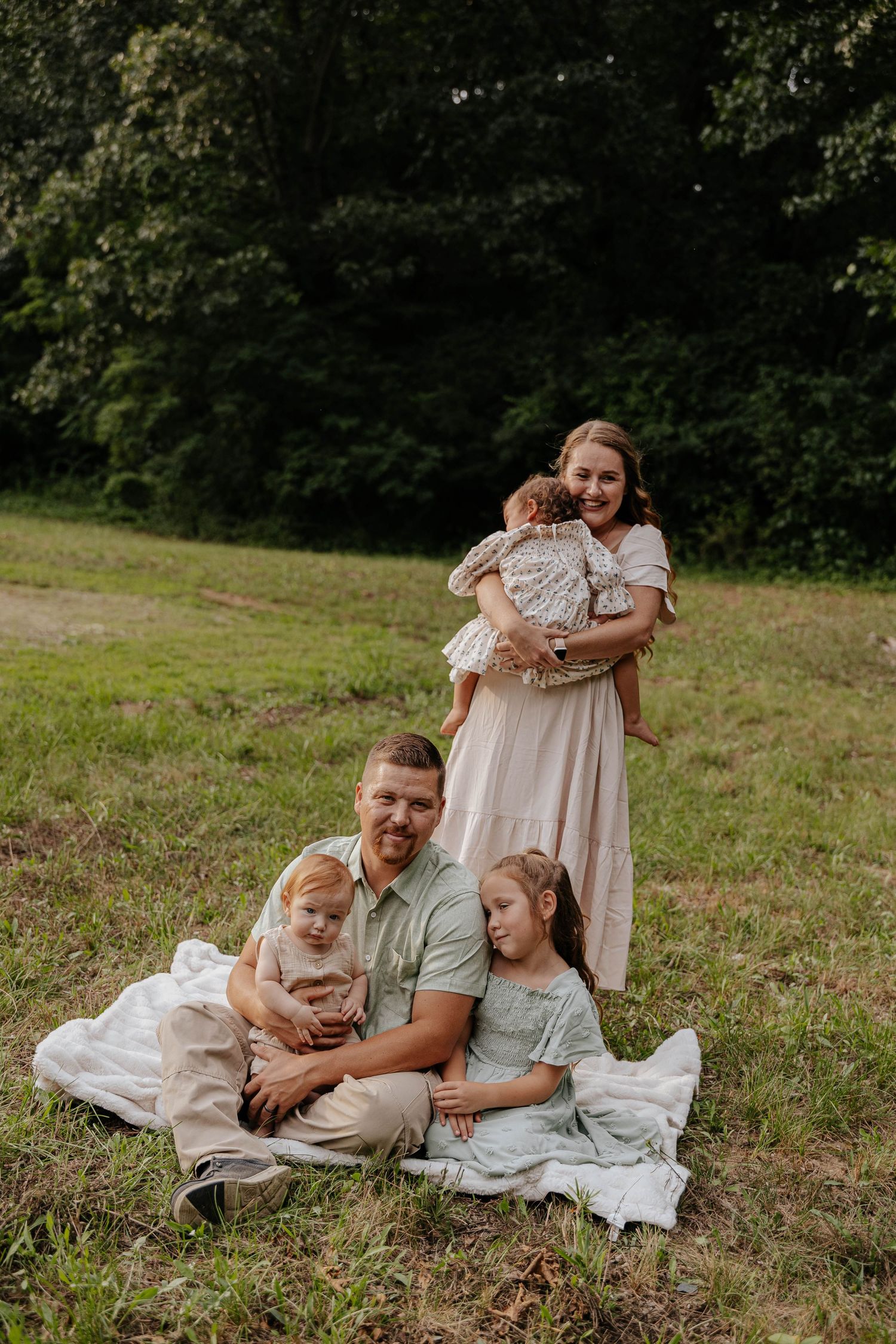 Outdoor family photography session featuring a mother standing with her baby while other family members sit together on a blanket in a grass