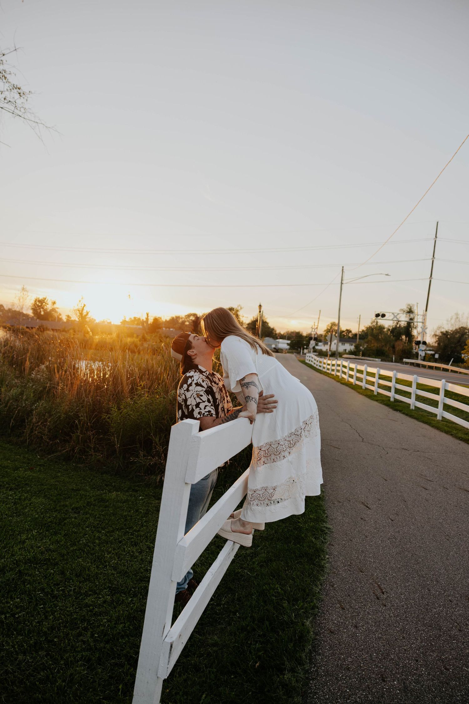 Engaged couple embraces beside a white fence at sunset during their Celery Flats engagement session in Portage, Michigan.