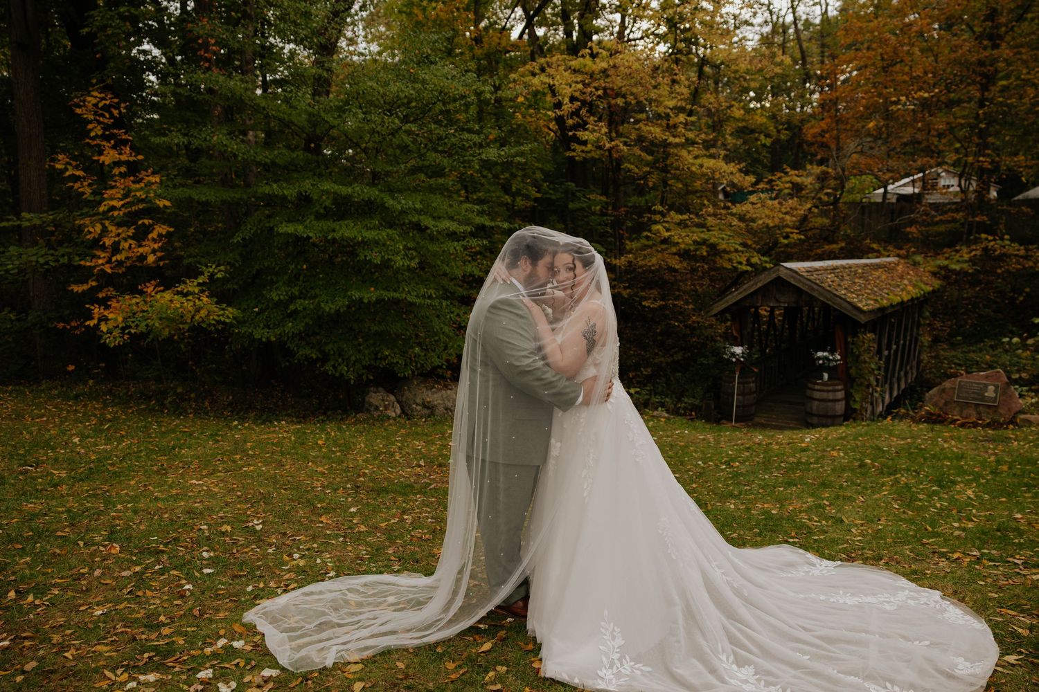 Bride & Groom poses in a flowing white wedding gown and long veil surrounded by autumn trees at Bowen Mills in Middleville, Michigan.