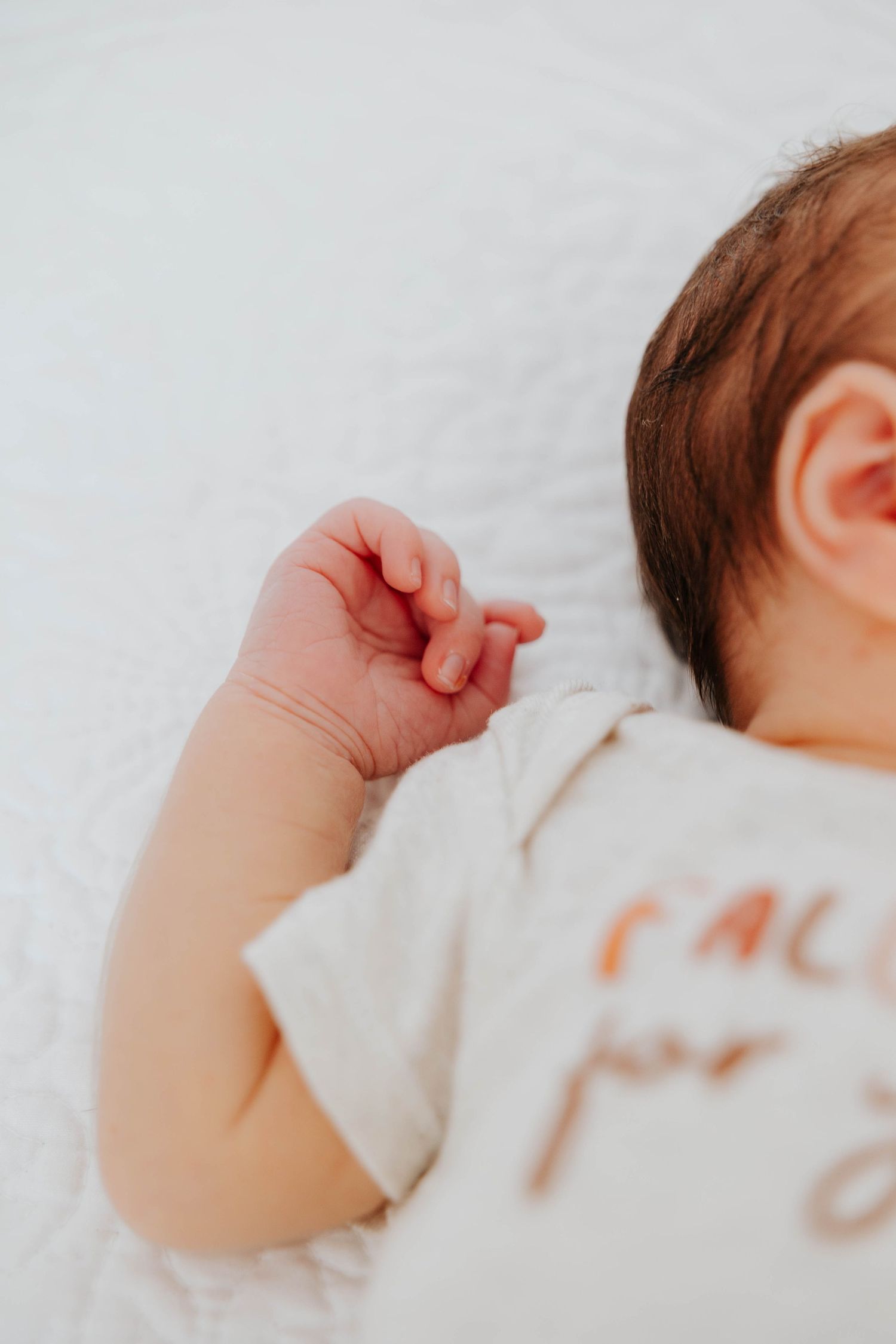 Close-up newborn photography capturing a baby's tiny raised hand, soft features, and personalized outfit against a simple white background.