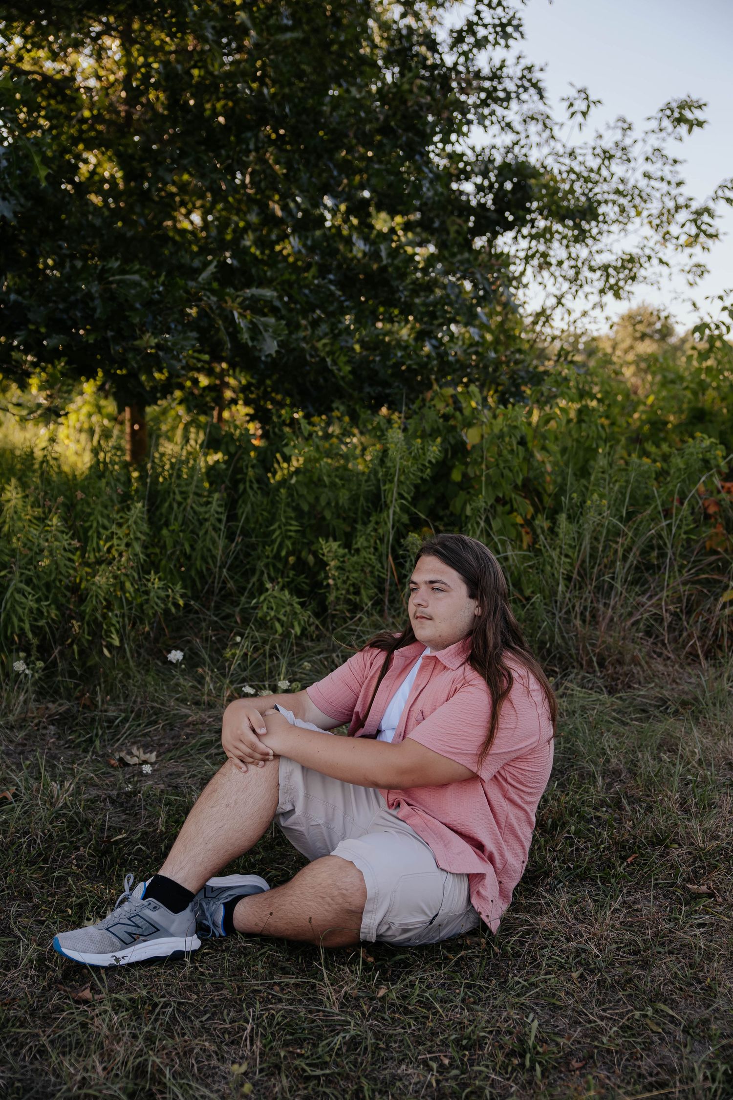 Outdoor senior portrait of a boy sitting in a grassy field surrounded by trees and greenery.