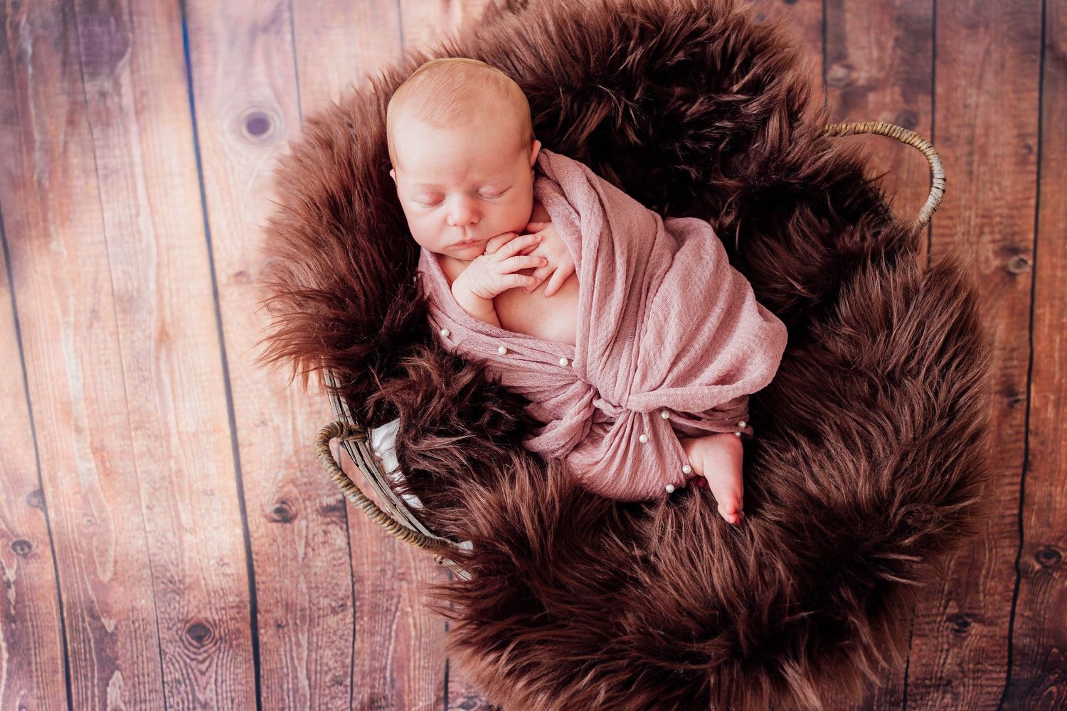 Posed newborn photography of a sleeping baby wrapped in a dusty pink blanket, nestled in a soft brown fur basket on a rustic wooden floor.
