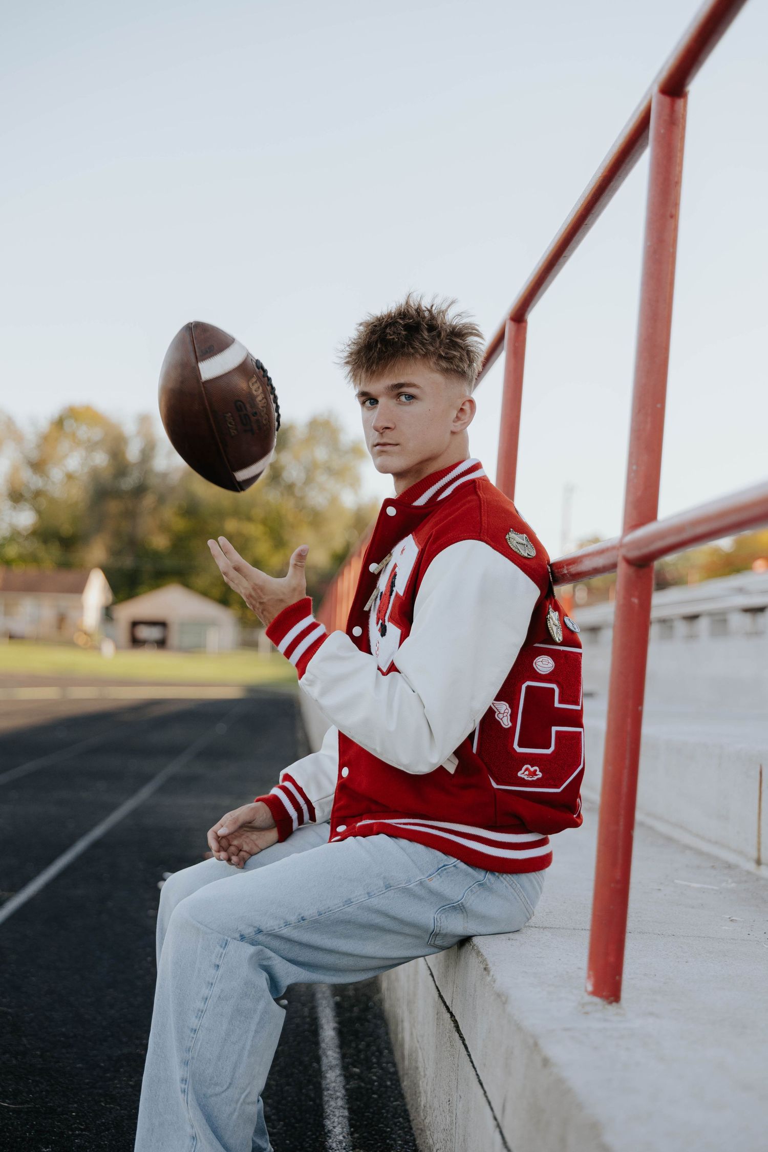 High school sports senior portrait featuring a football player in a red and white varsity jersey, sitting on outdoor bleachers.