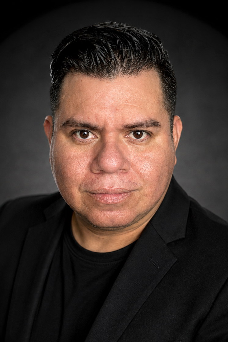 Professional headshot of a man in a black blazer with dark hair against a gray background.