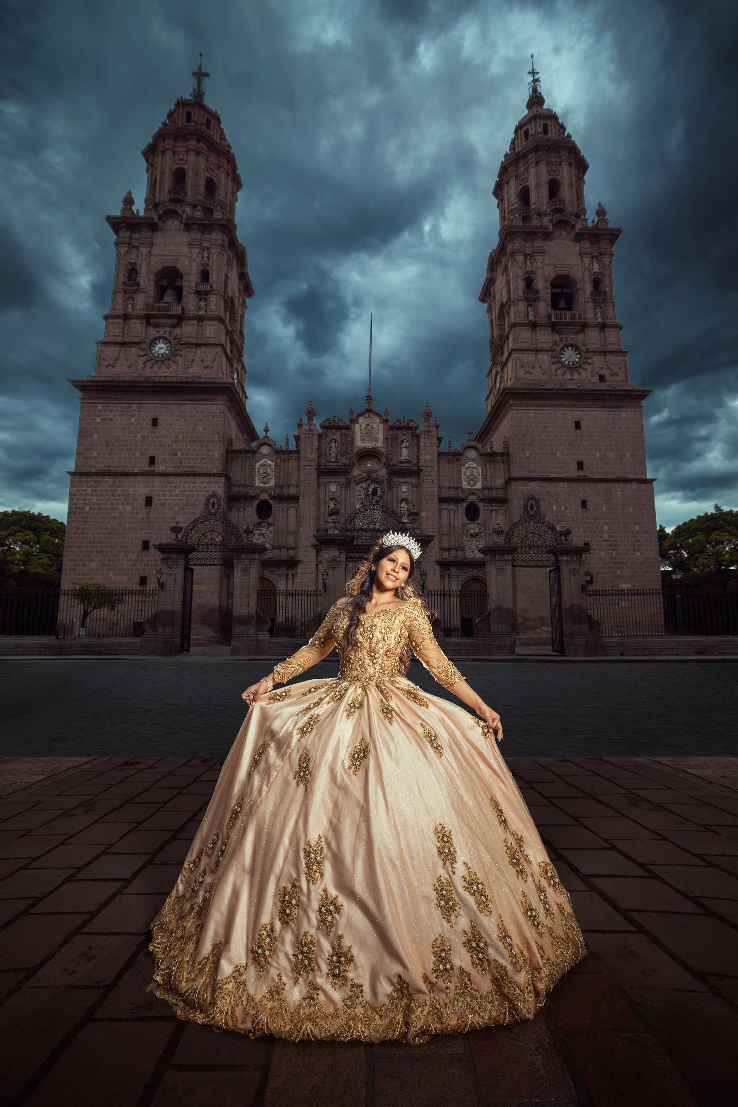 Young woman in gold quinceañera gown poses before a grand cathedral under dramatic stormy skies.
