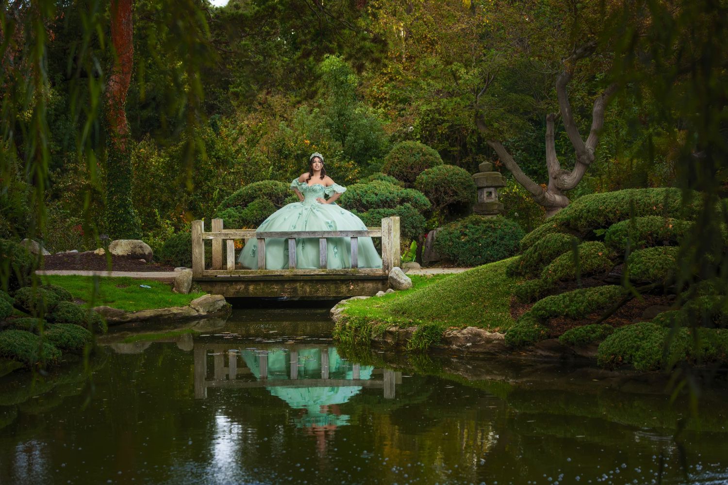 Woman in mint green ball gown poses on bridge over pond in lush Japanese garden.