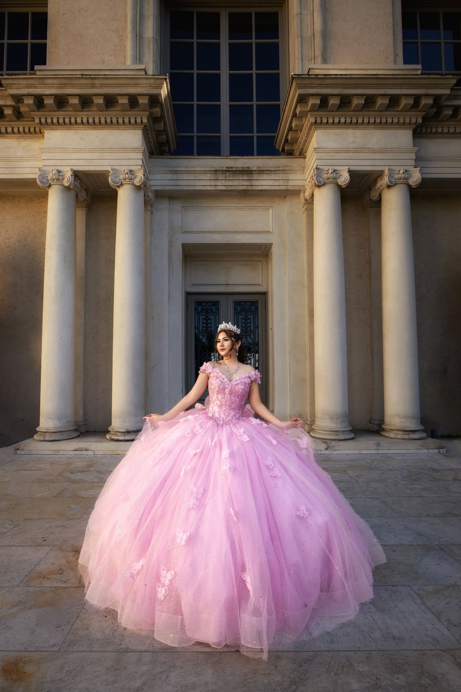 Young woman in a voluminous pink ball gown with floral details and tiara poses before grand columns.