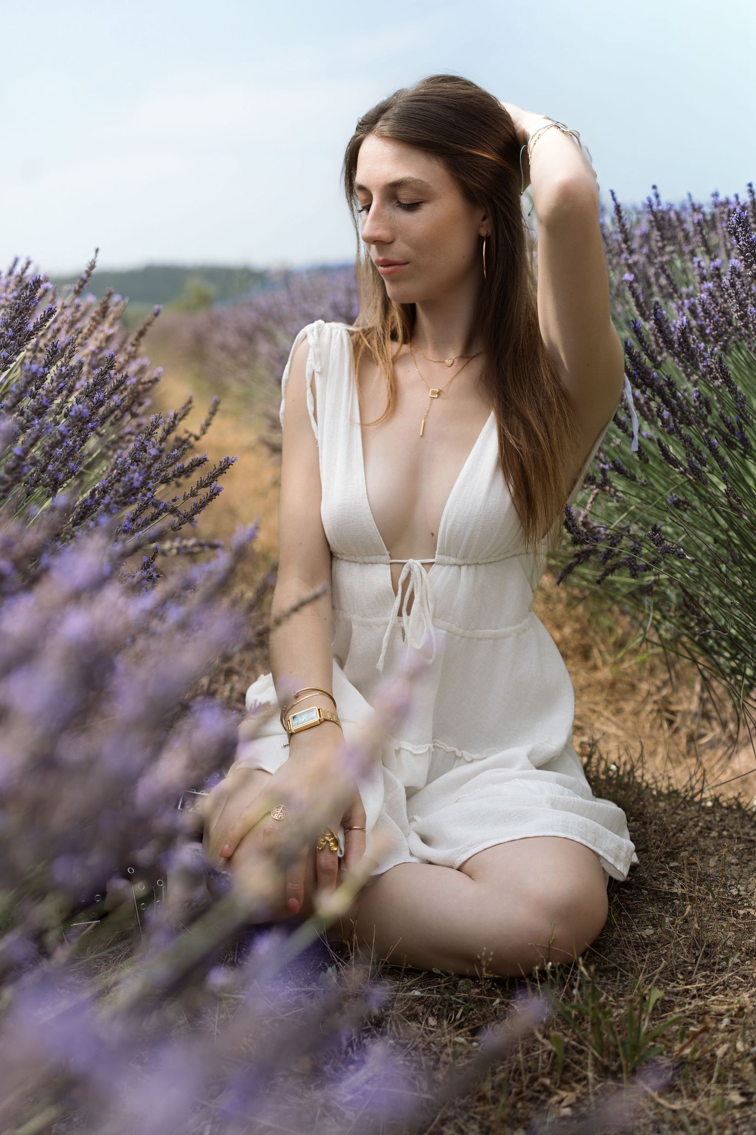 Woman in white dress sitting in lavender field, wearing gold jewelry, with hand in hair.