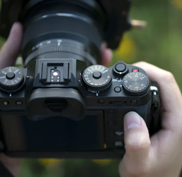 Gros plan de mains tenant un appareil photo sans miroir Fujifilm avec un grand objectif, à l'extérieur avec un fond de feuillage vert.