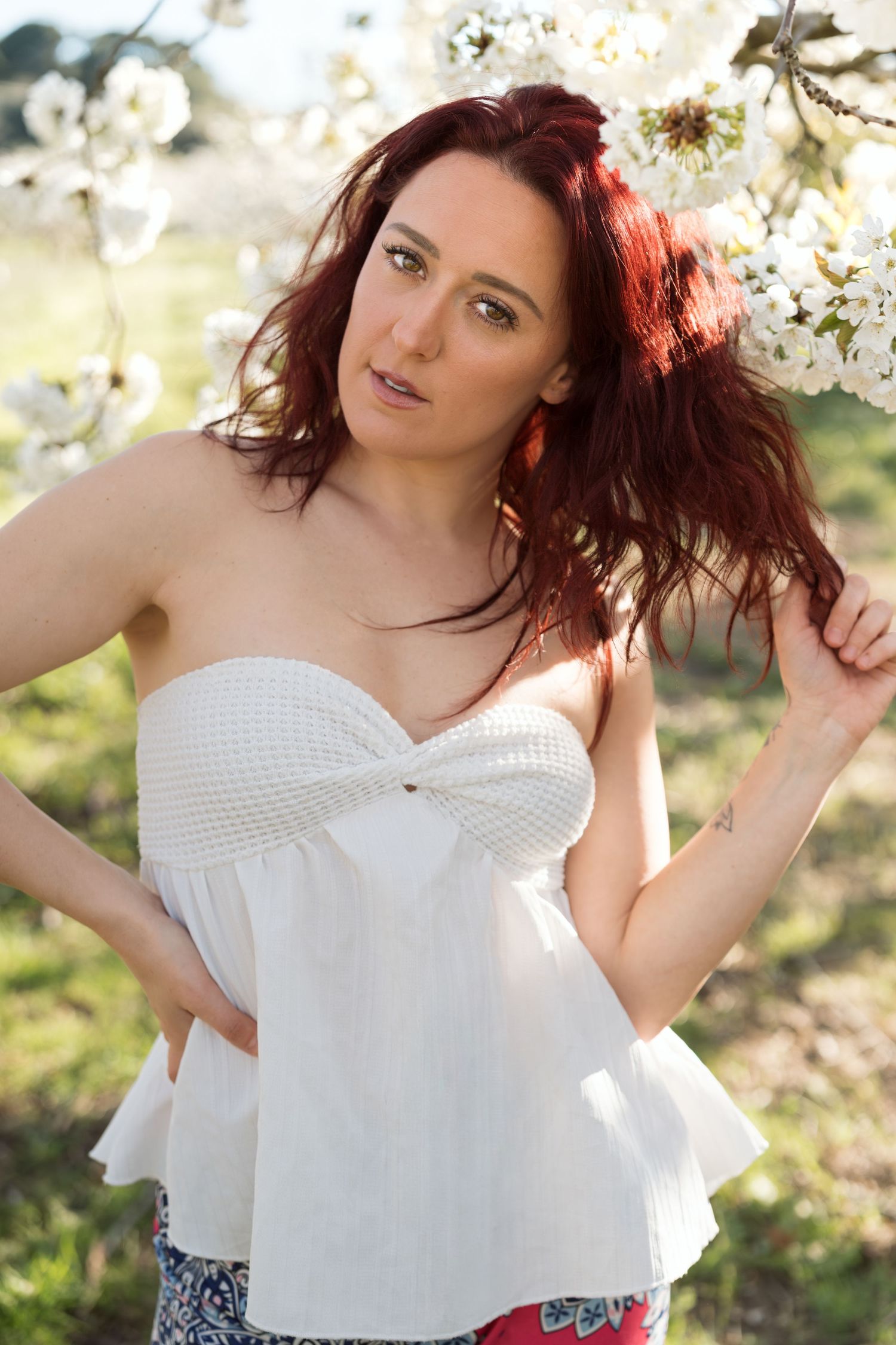 A woman with red hair poses in a white strapless top among blooming white cherry blossom trees outdoors.