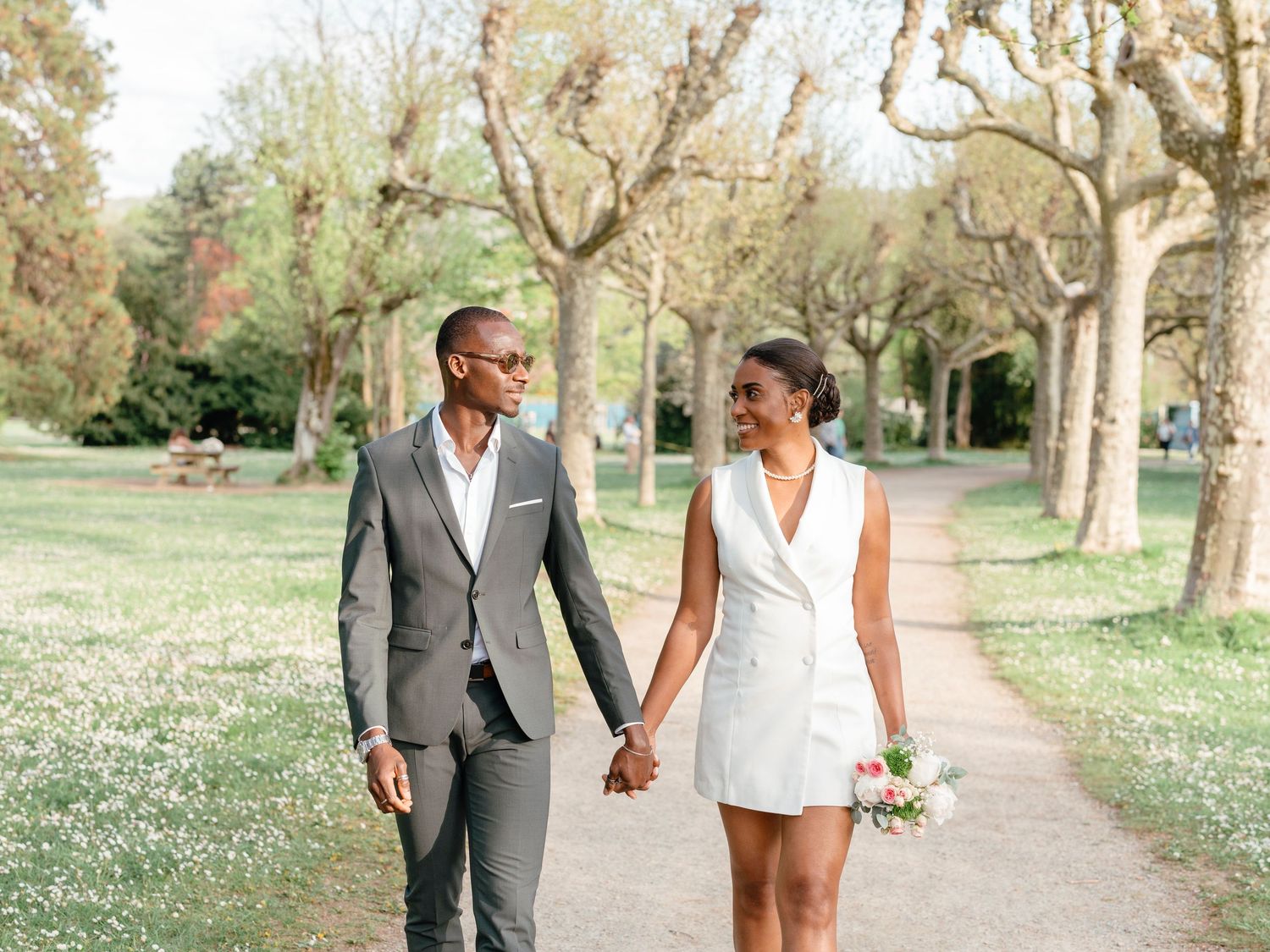 Joyful newlywed couple holding hands and sharing a smile while walking through a sunlit park.