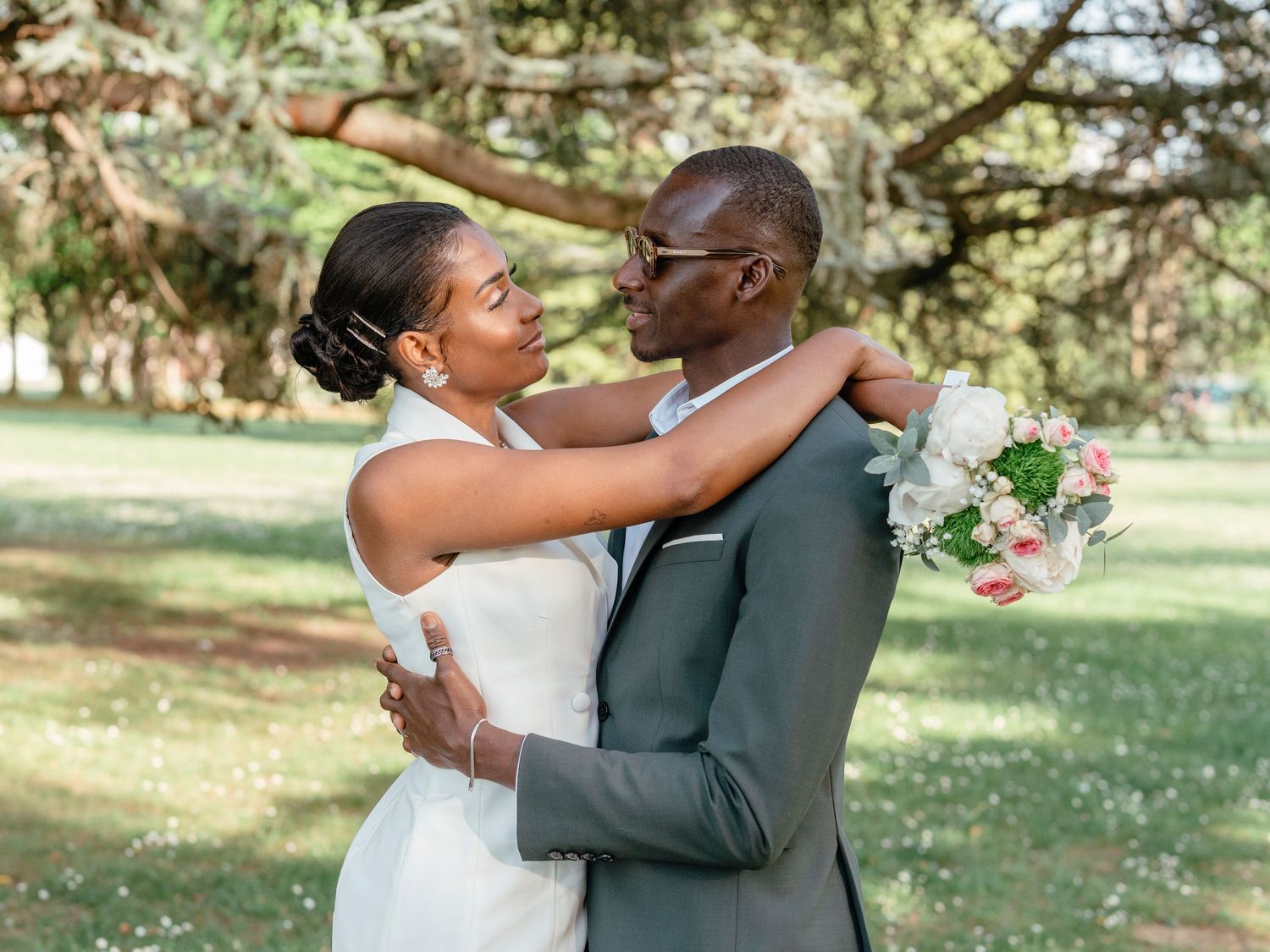 Newlywed couple embracing and smiling at each other in a lush green park, groom holding a pink and white bouquet.