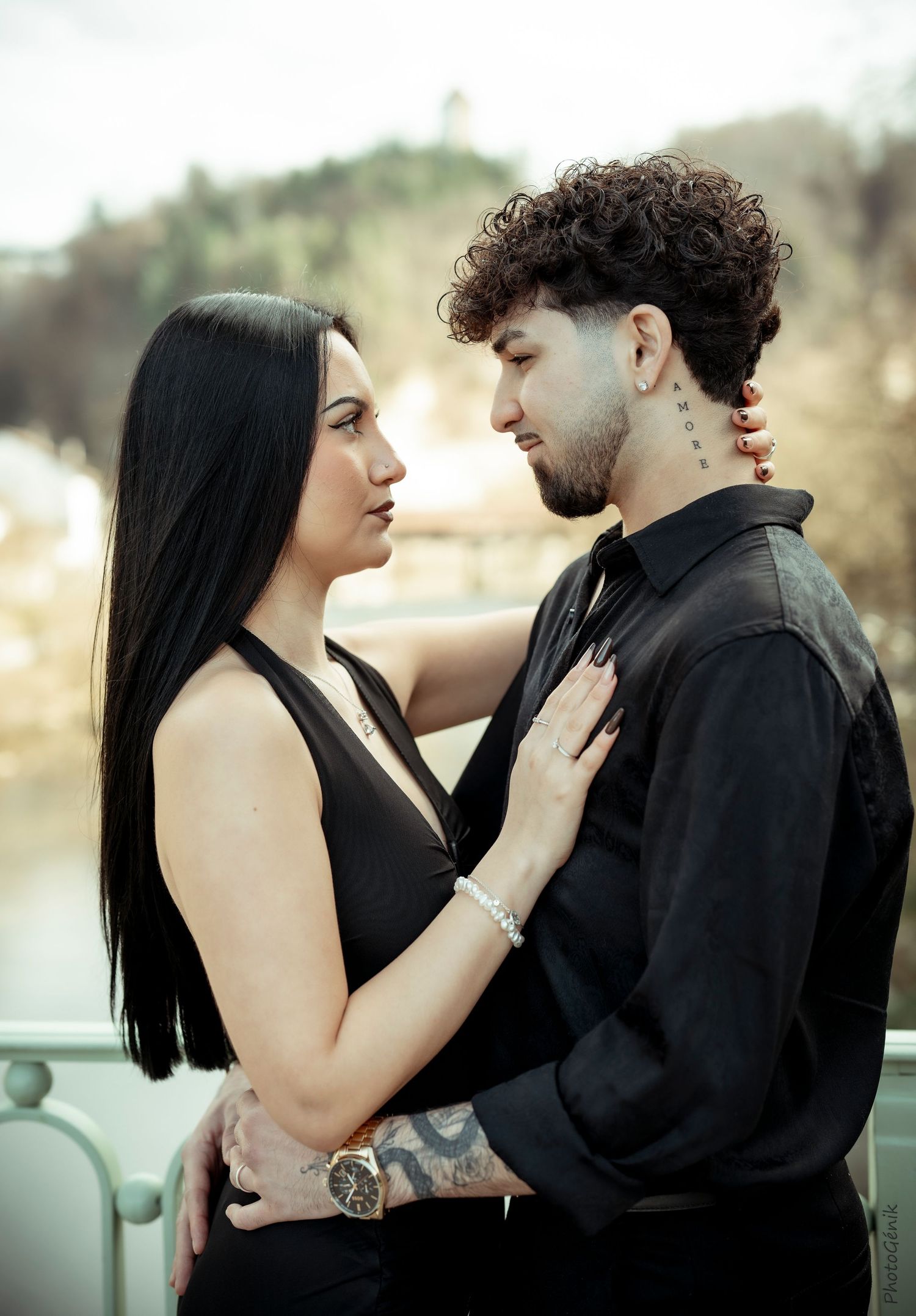 Couple dressed in black embrace intimately on a bridge with blurred outdoor background behind them.