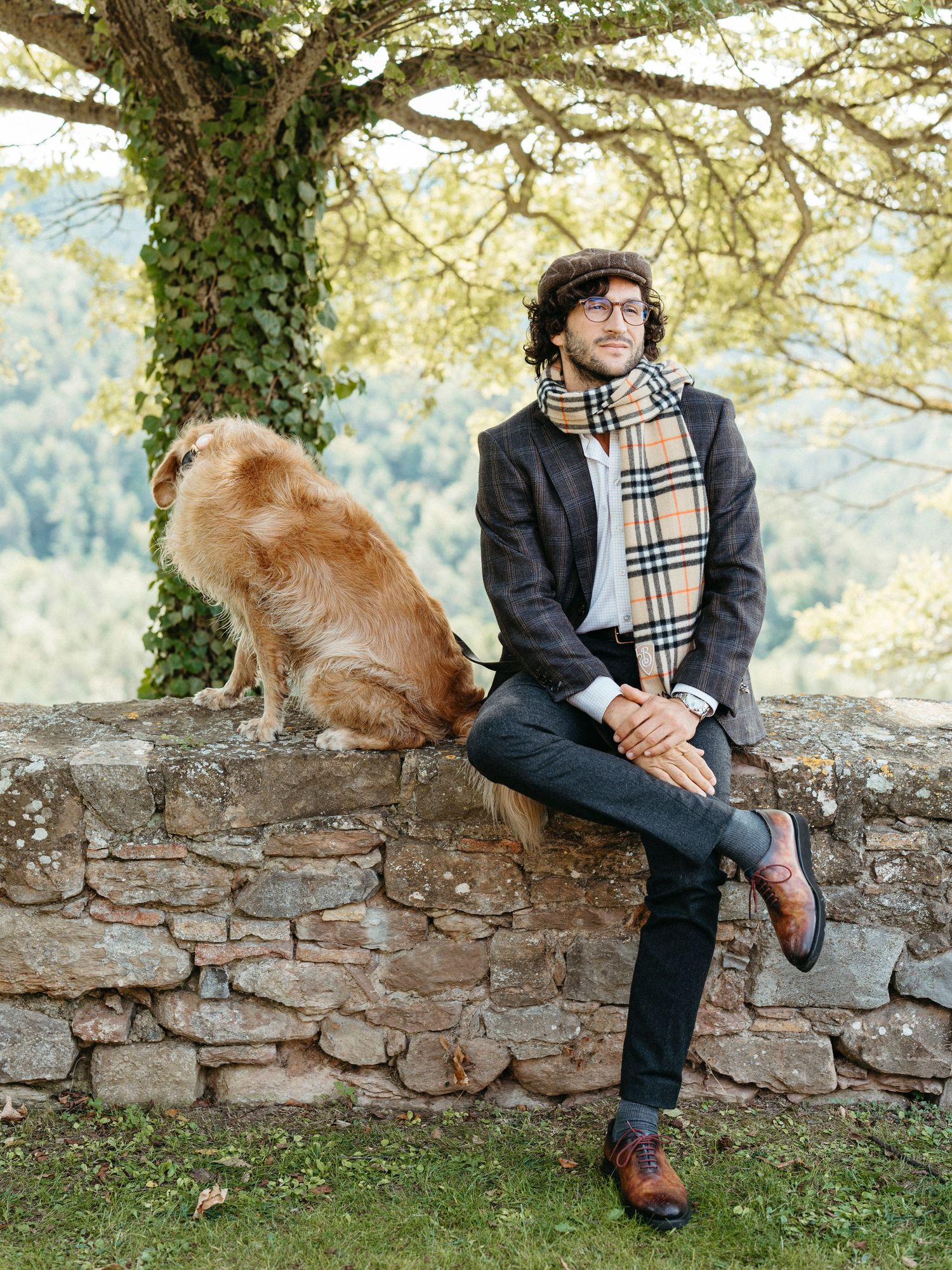 Dapper man in flat cap and checkered scarf sits on stone wall beside golden retriever under a tree.