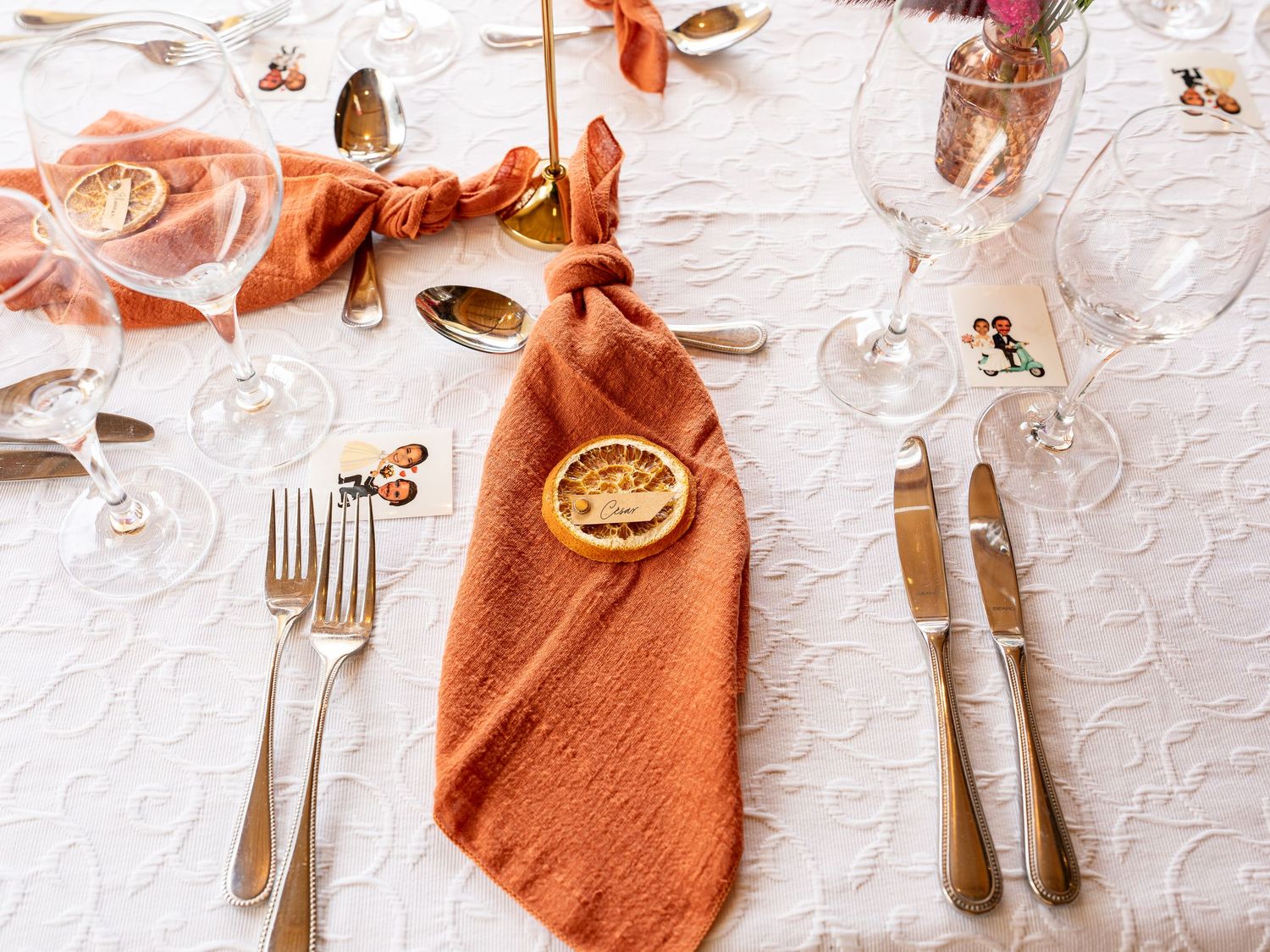 Elegant wedding table setting with terracotta linen napkin, gold cutlery, crystal glasses, and decorative place card.