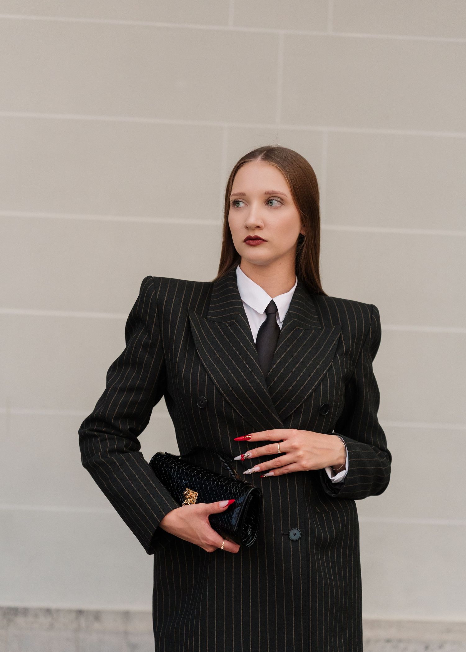 Woman in black pinstripe suit with white shirt and tie, holding a clutch bag, posing against a gray wall.