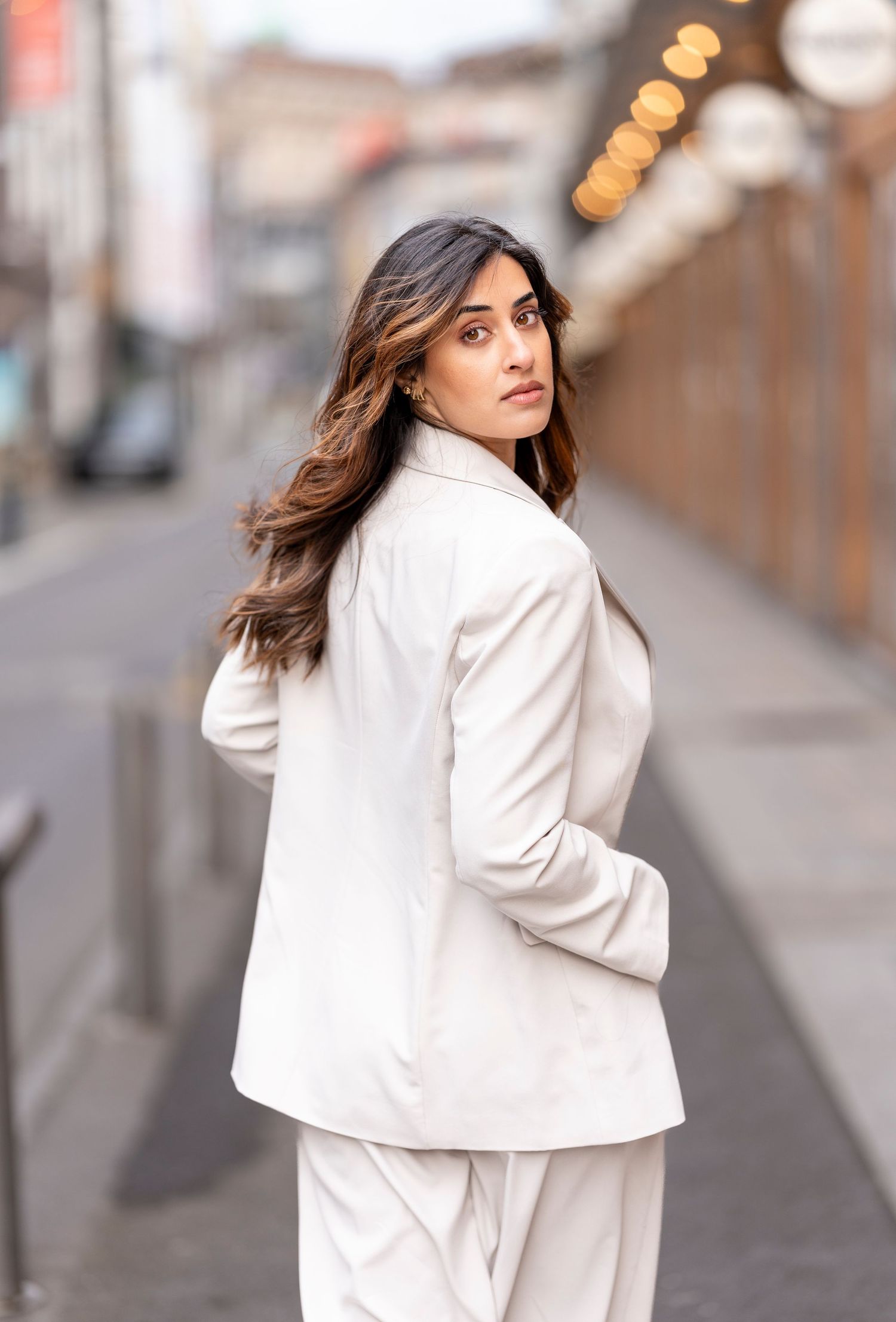 Woman in cream double-breasted blazer and matching trousers poses on urban street looking over shoulder.