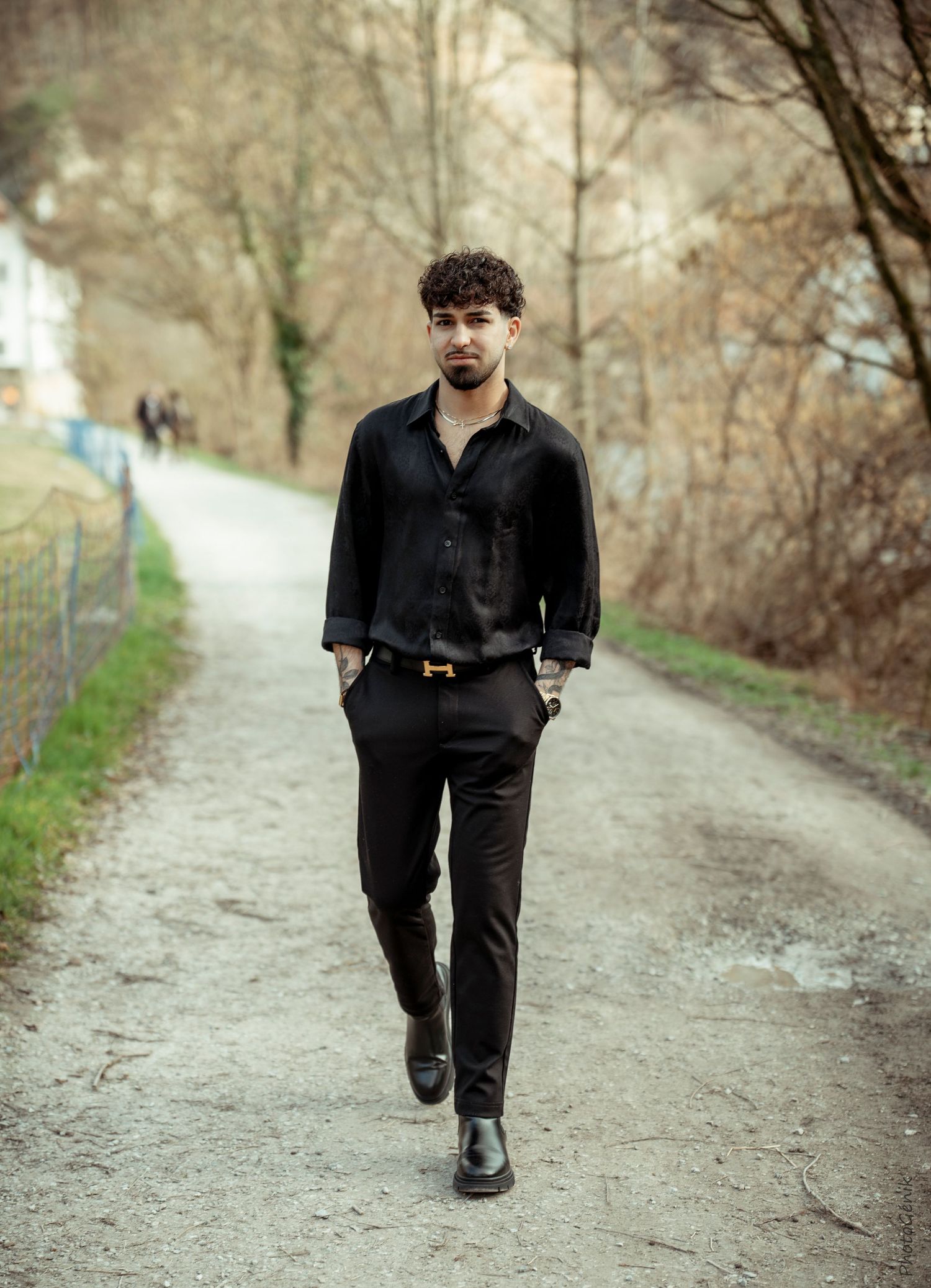 Stylish young man in all-black outfit with gold belt walking confidently down a tree-lined pathway.