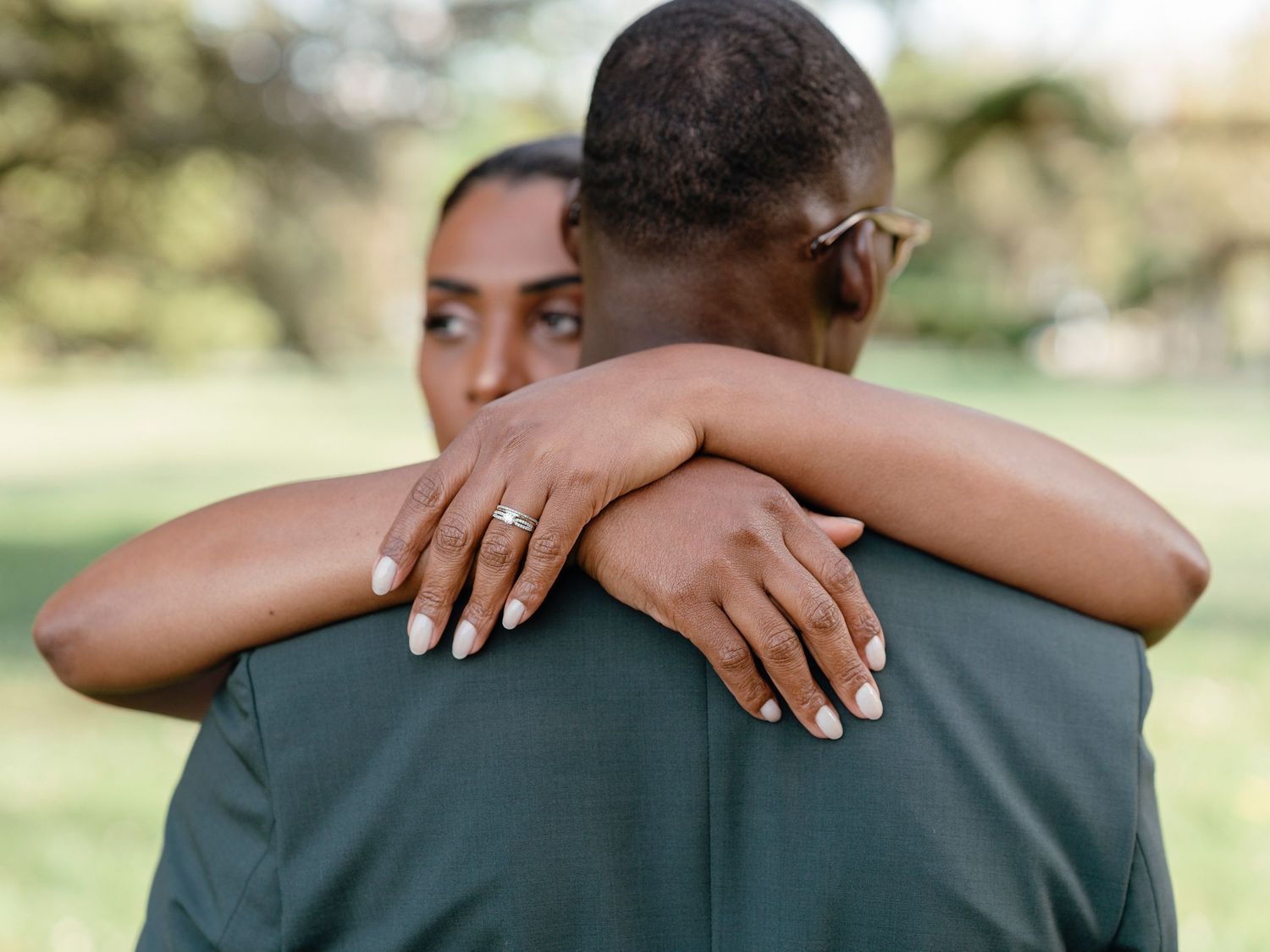 Close-up of bride embracing groom from behind, highlighting her diamond ring and manicured nails against his gray suit.