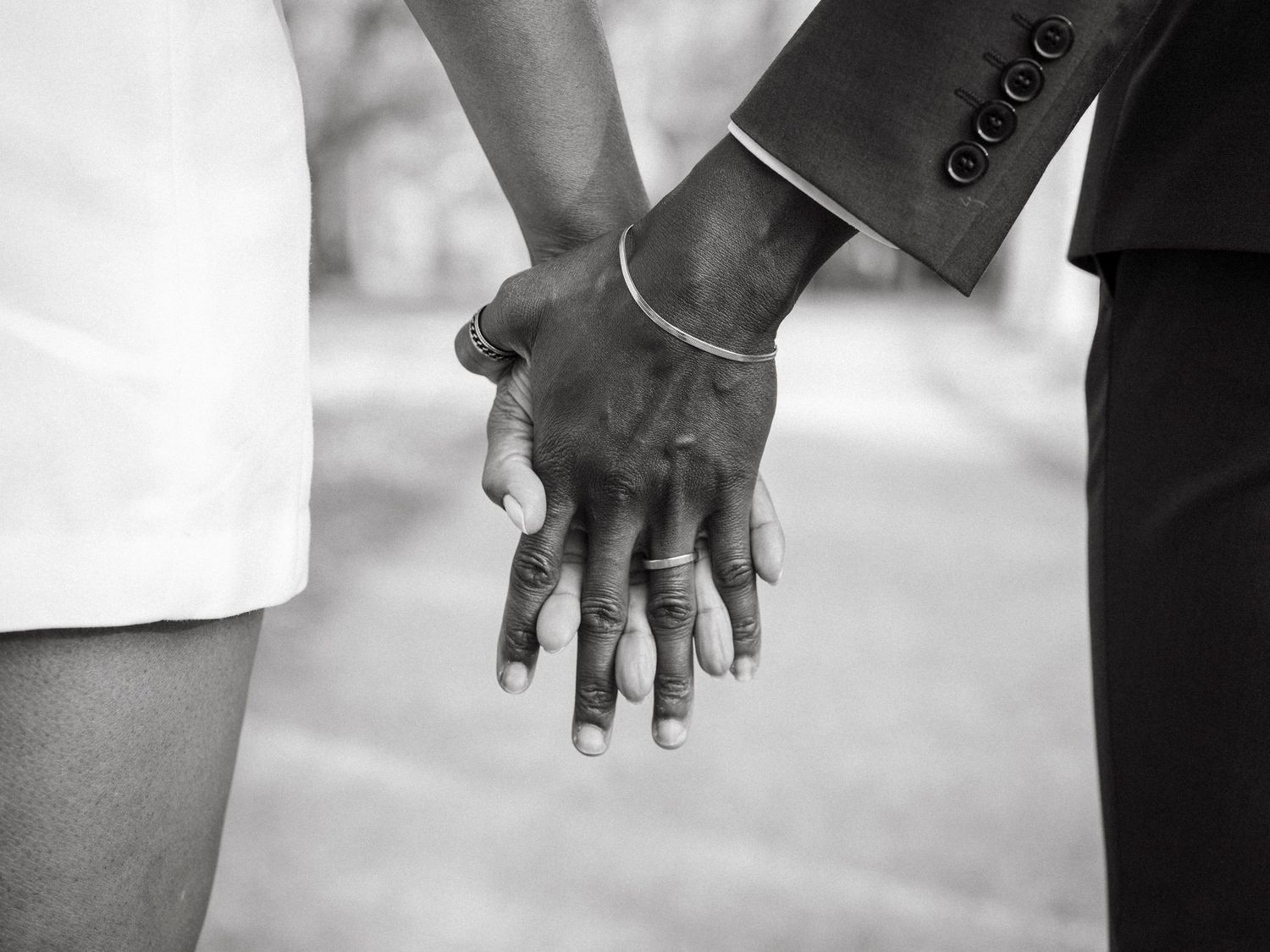 Black and white close-up of newlyweds holding hands, showcasing wedding bracelet and rings.