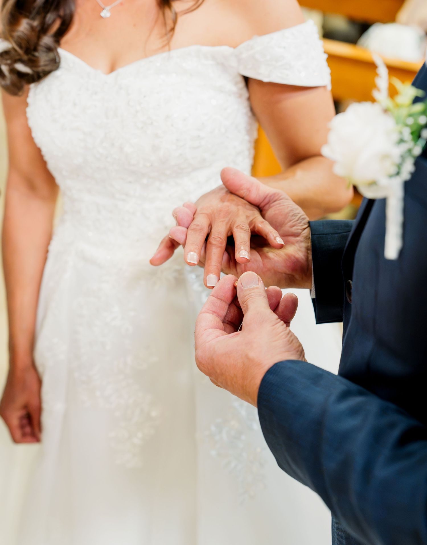 Groom placing a wedding ring on the bride's finger during a ceremony, with the bride in a white off-shoulder gown.