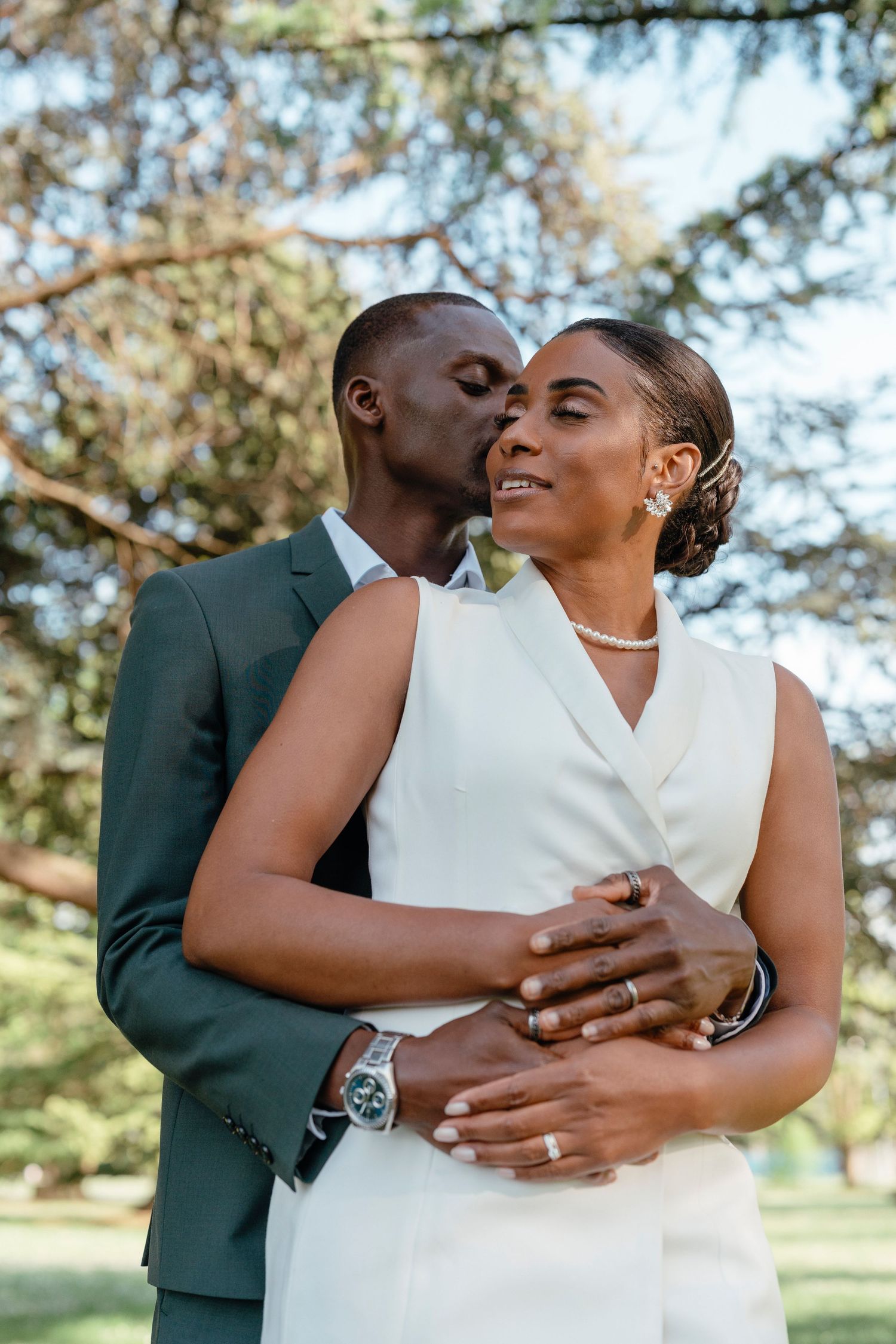 A groom kisses his bride on the cheek as she smiles, both dressed in elegant wedding attire outdoors.