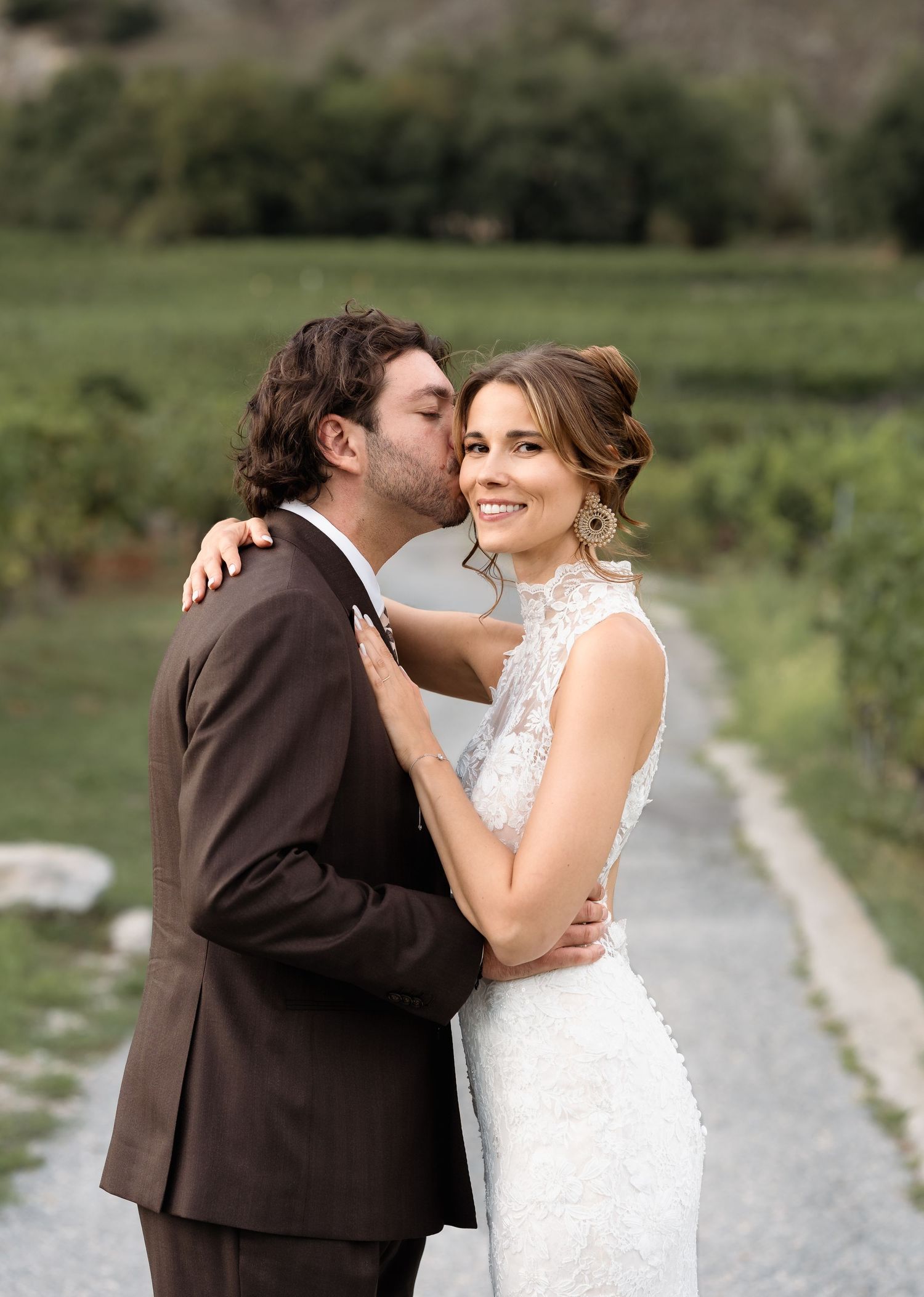 A bride and groom share a romantic kiss in a lush vineyard setting.