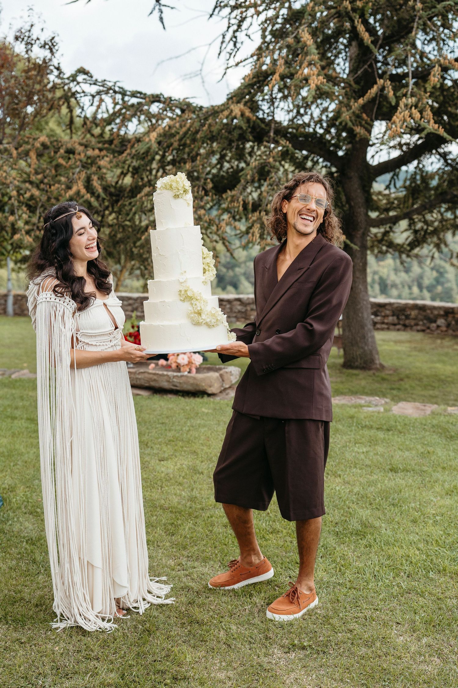 Two brides laughing while holding a white tiered wedding cake outdoors on a green lawn.
