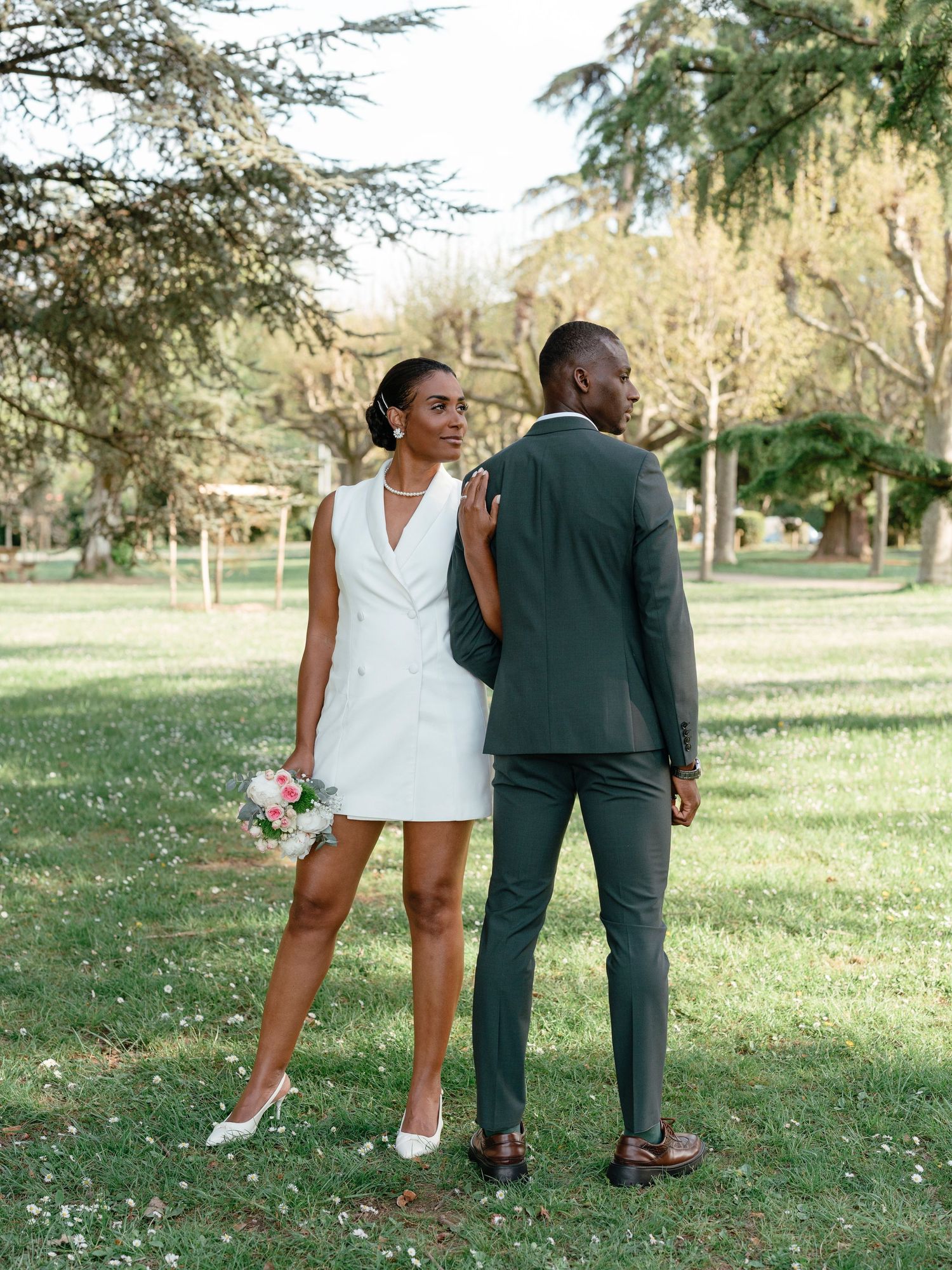 A bride in a white mini dress holds a bouquet while standing back-to-back with her groom in a park.