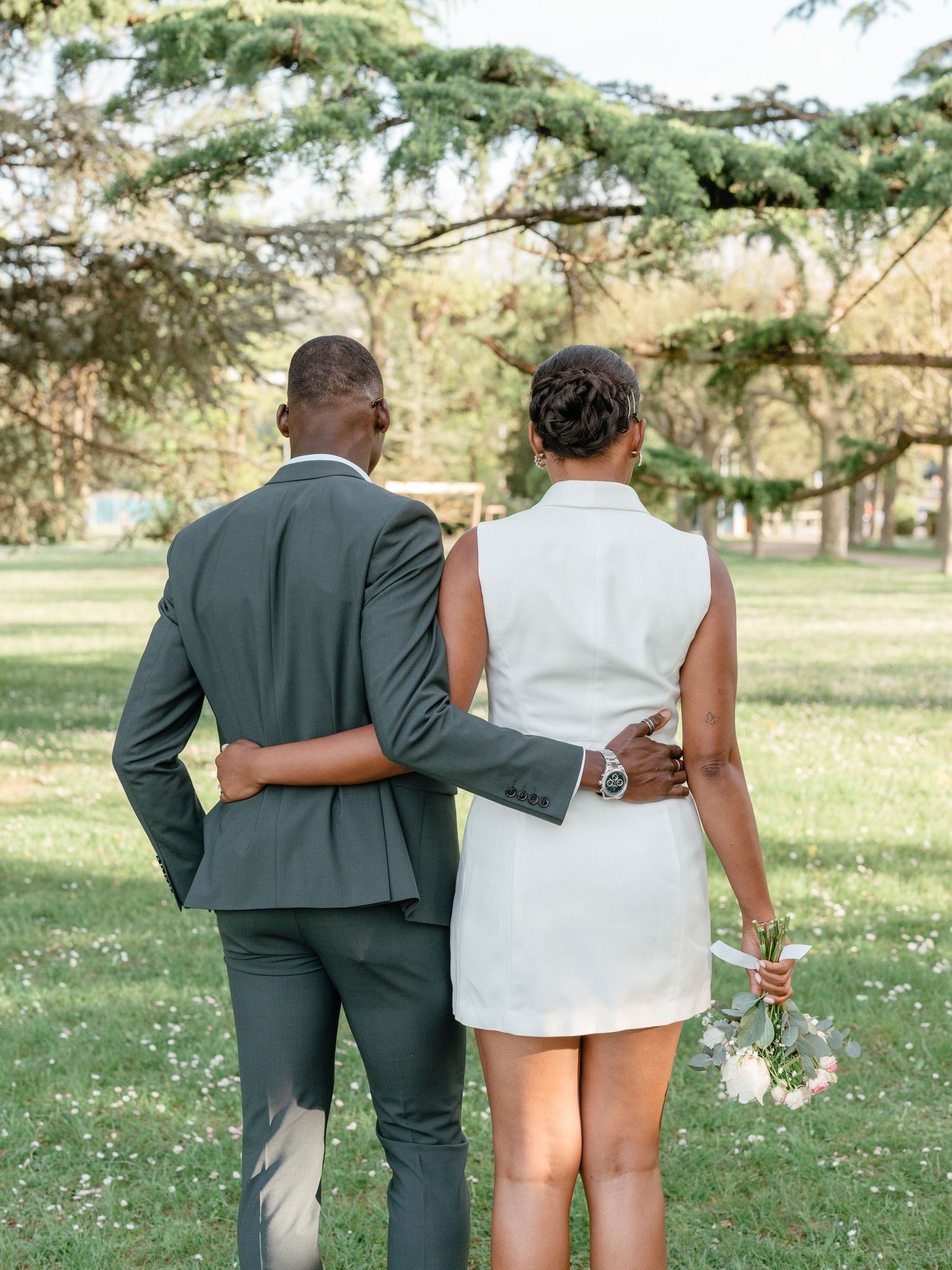 Newlywed couple walking hand in hand through lush green park, bride in white mini dress holding bouquet.