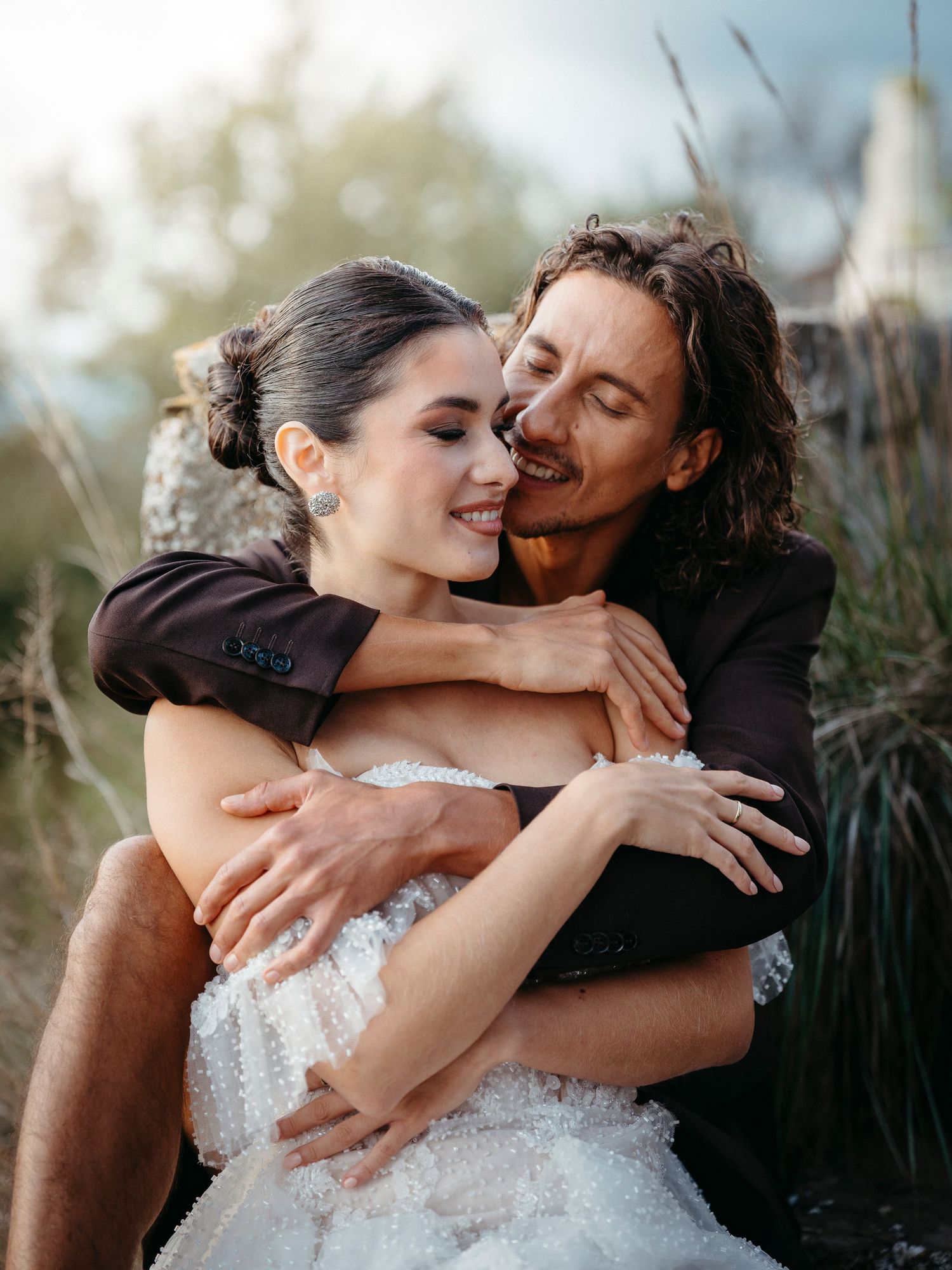 Joyful wedding couple embracing outdoors, bride in white lace gown with groom in dark suit.