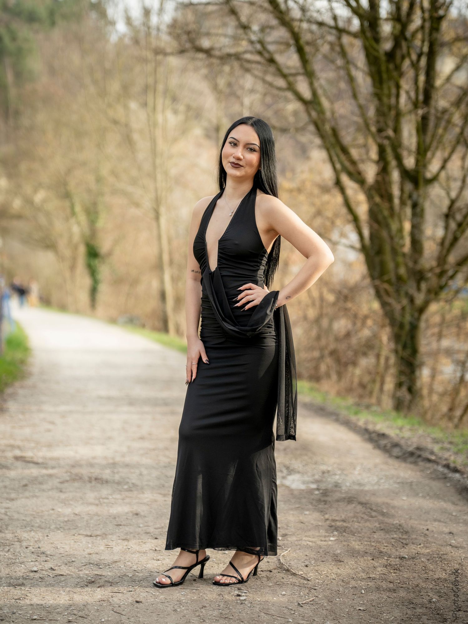 Elegant woman in a black halter maxi dress posing on a tree-lined gravel path outdoors.
