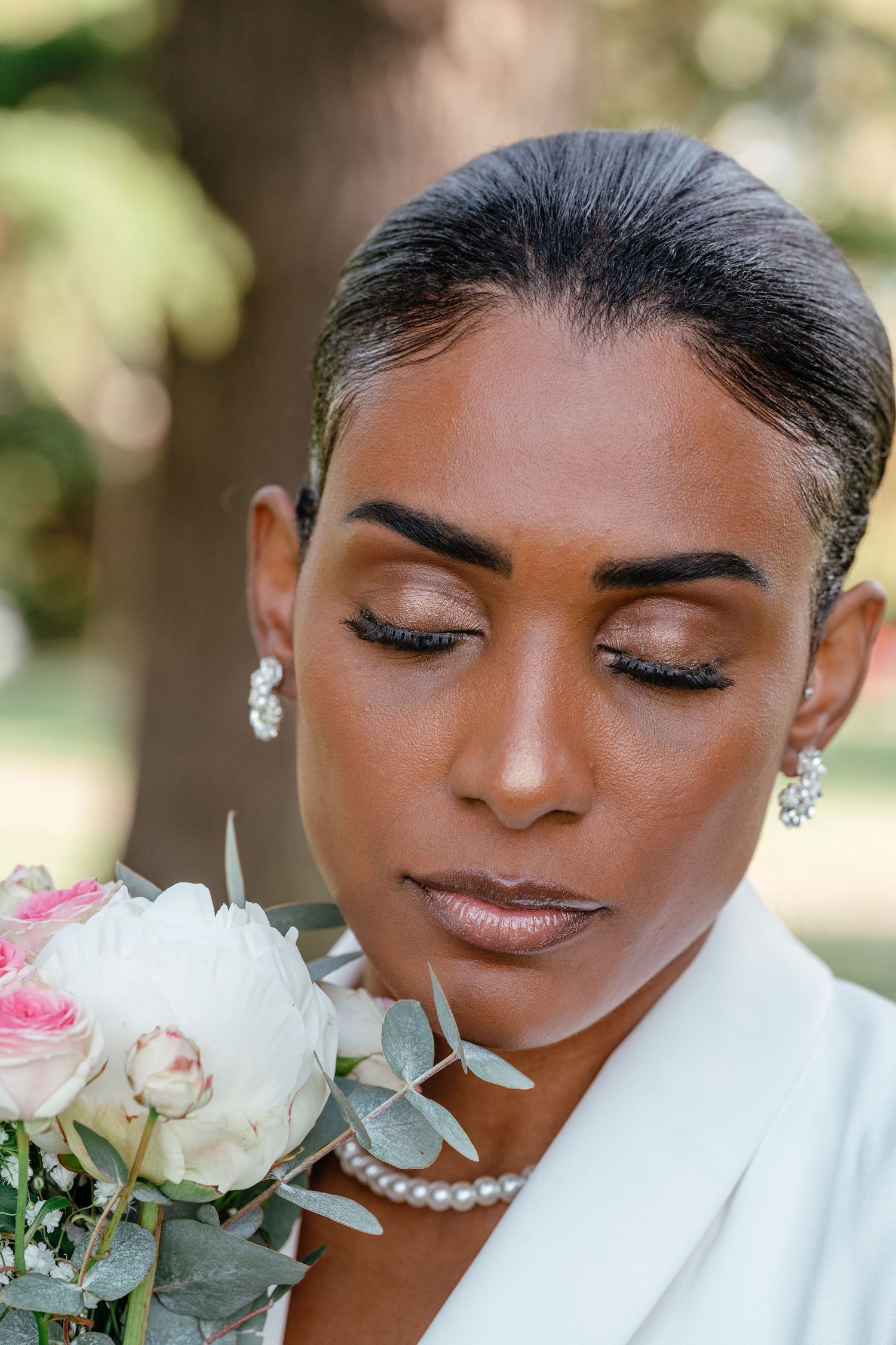 Elegant Black bride with closed eyes holding pink and white roses, wearing pearl necklace and crystal earrings.