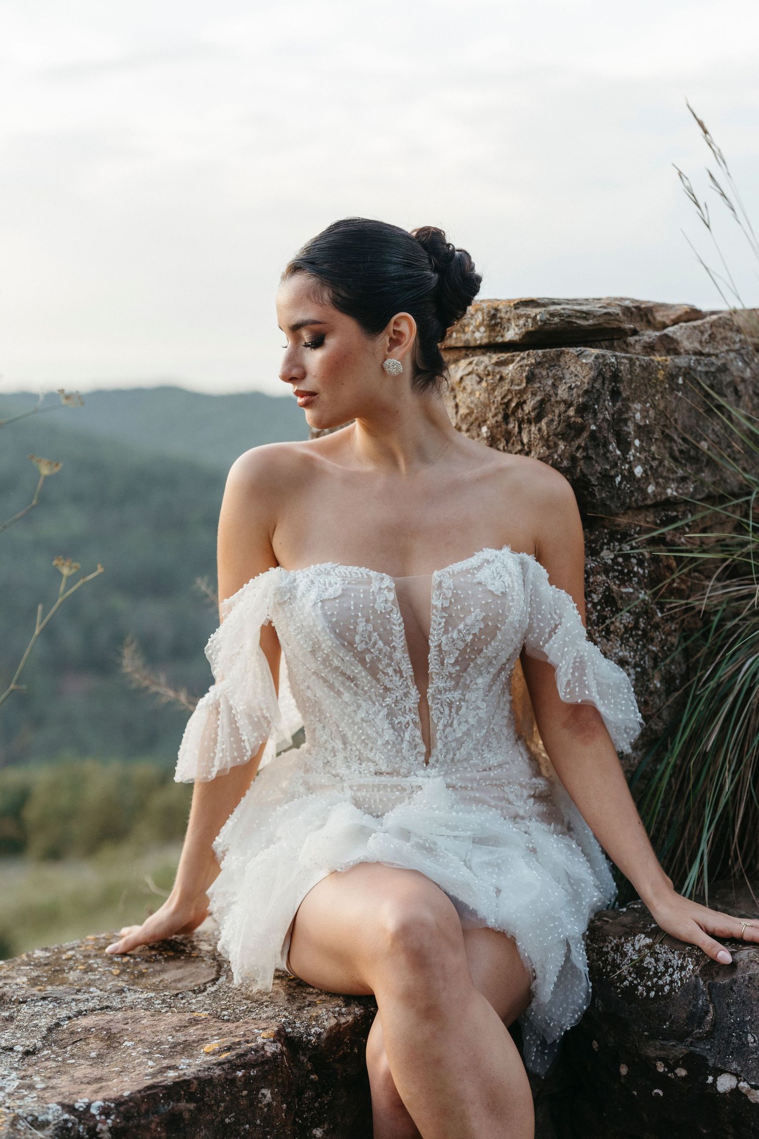 Bride in short off-shoulder lace wedding dress sits on stone wall with mountain views behind her.