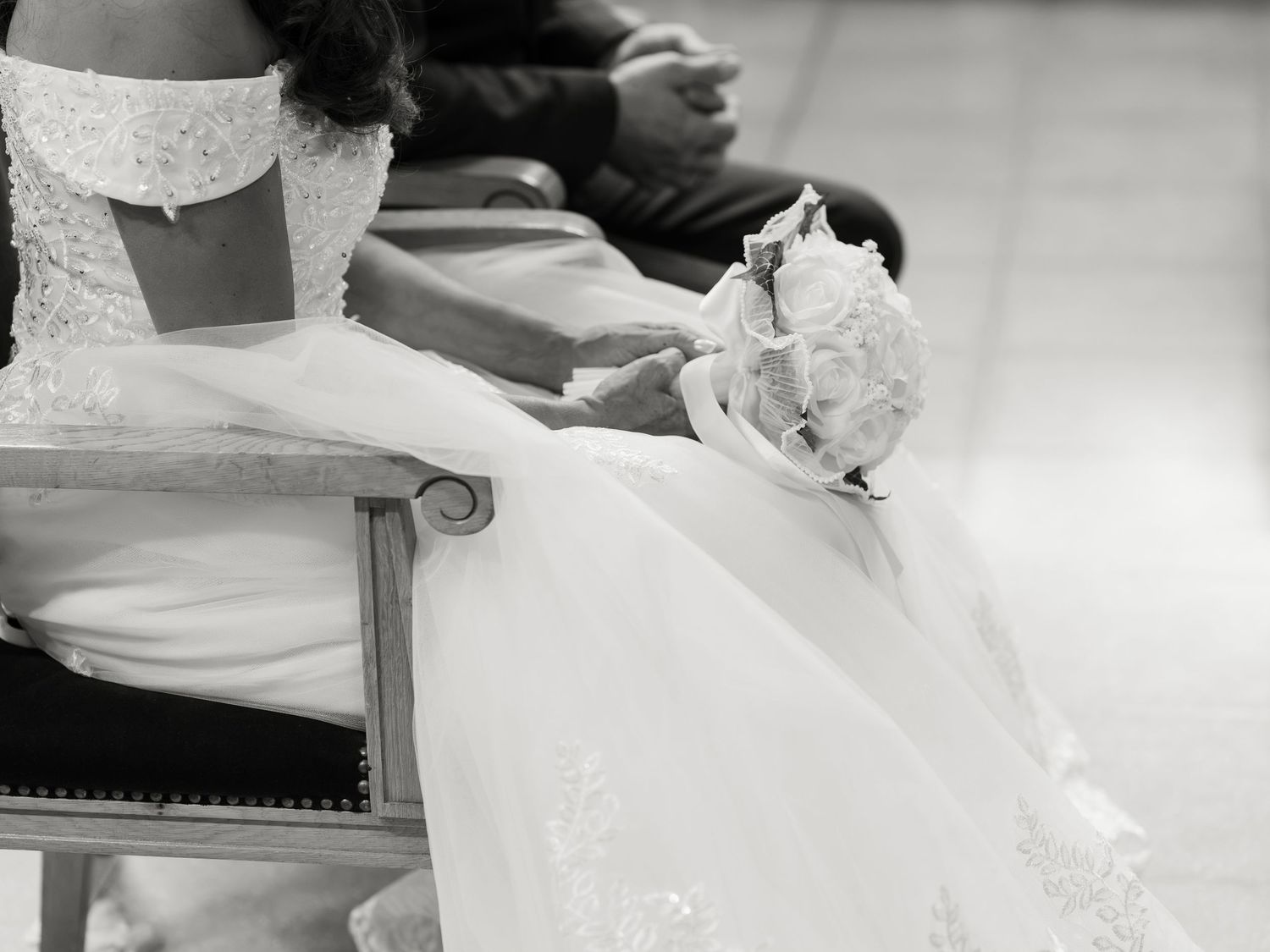 Black and white photo of a bride in a white lace gown holding a bouquet while seated at a wedding ceremony.