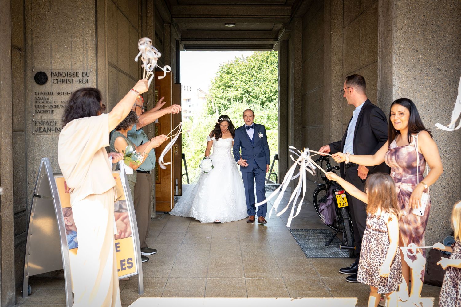Bride and groom exit ceremony venue through ribbon arch, greeted by guests celebrating with streamers outdoors.