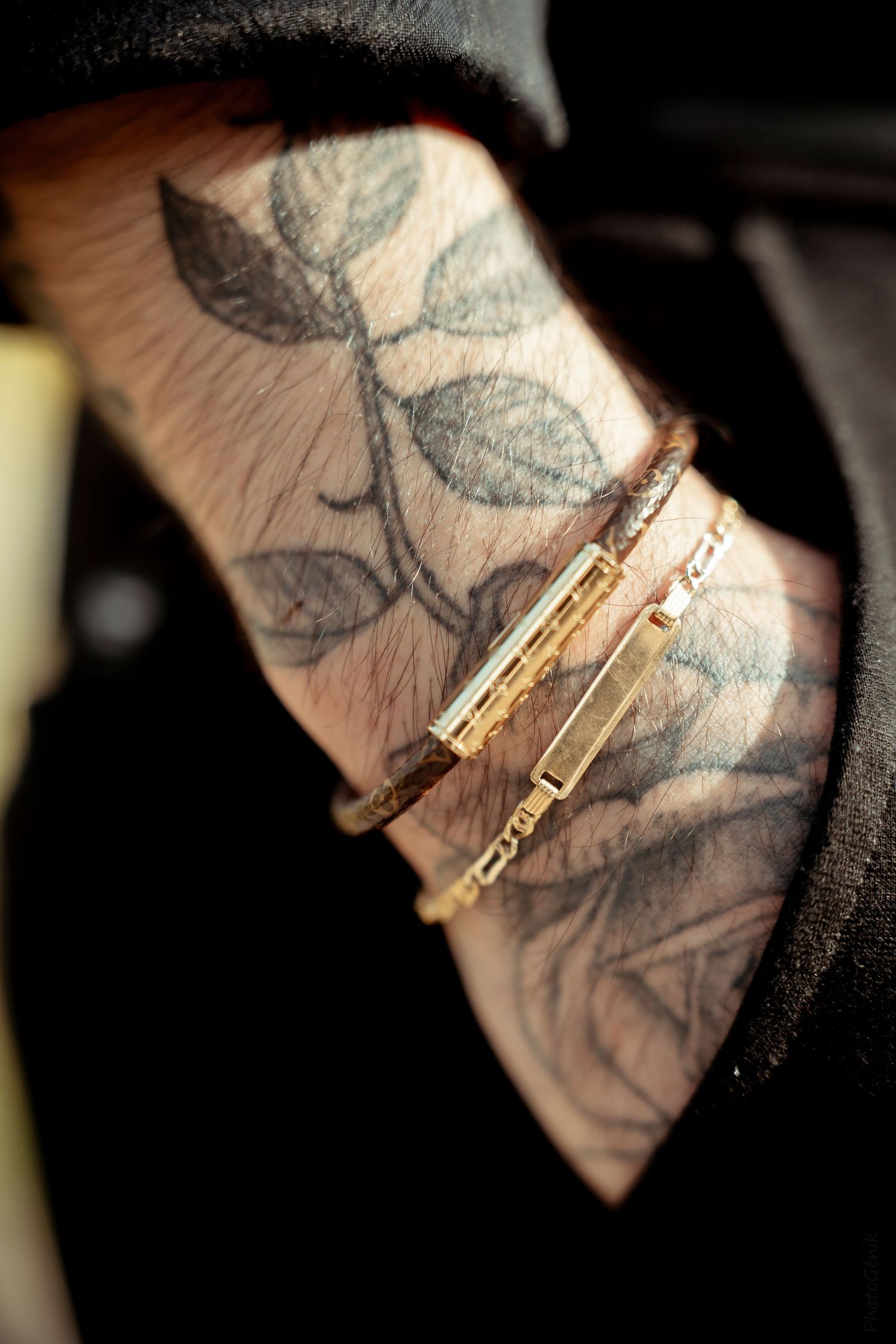 Close-up of a tattooed hand featuring floral leaf tattoos and gold bracelets against a dark background.